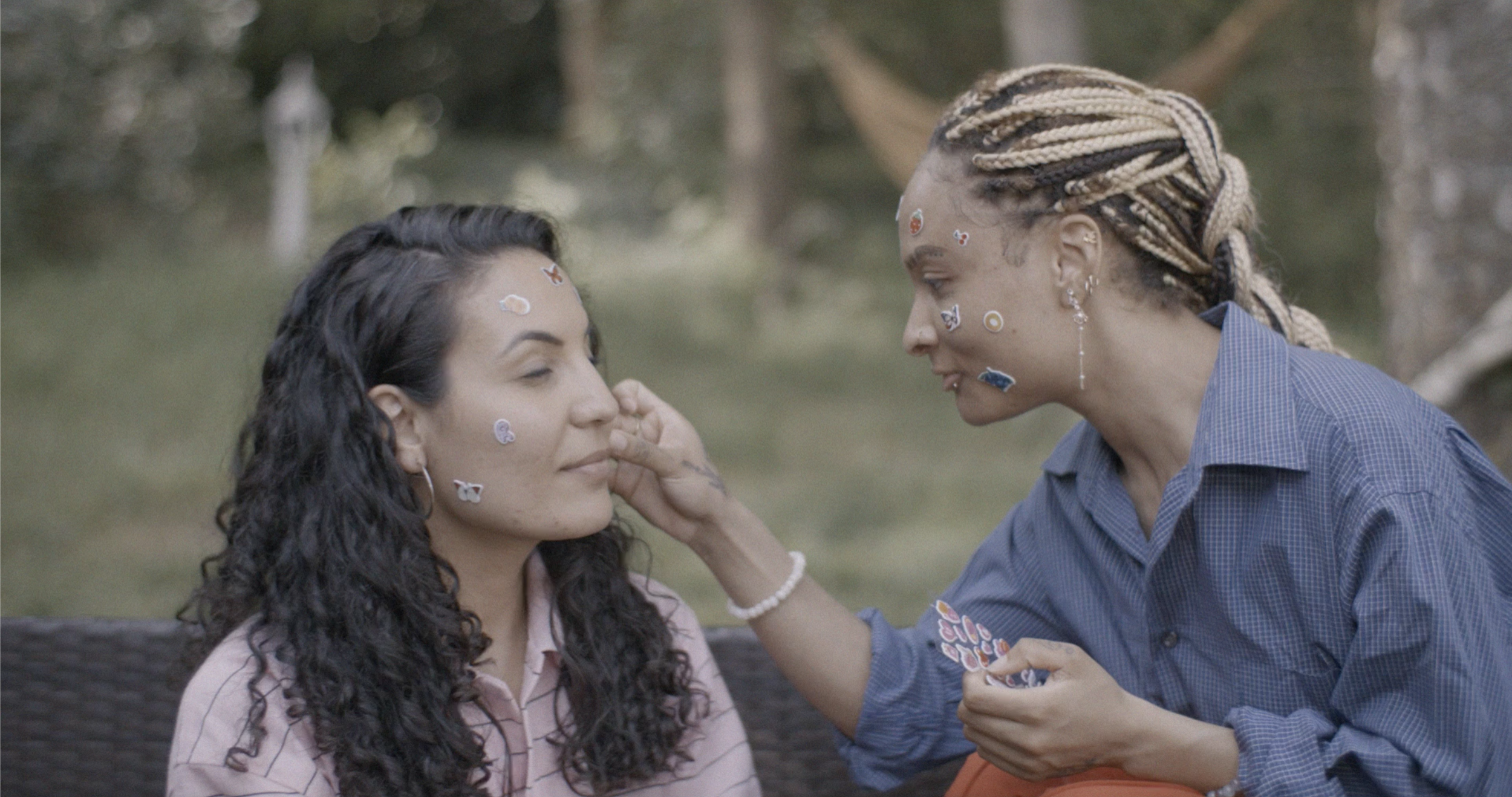 Two women with stickers on their faces and hair interacting outdoors, one touching the other's face, with trees in the background.