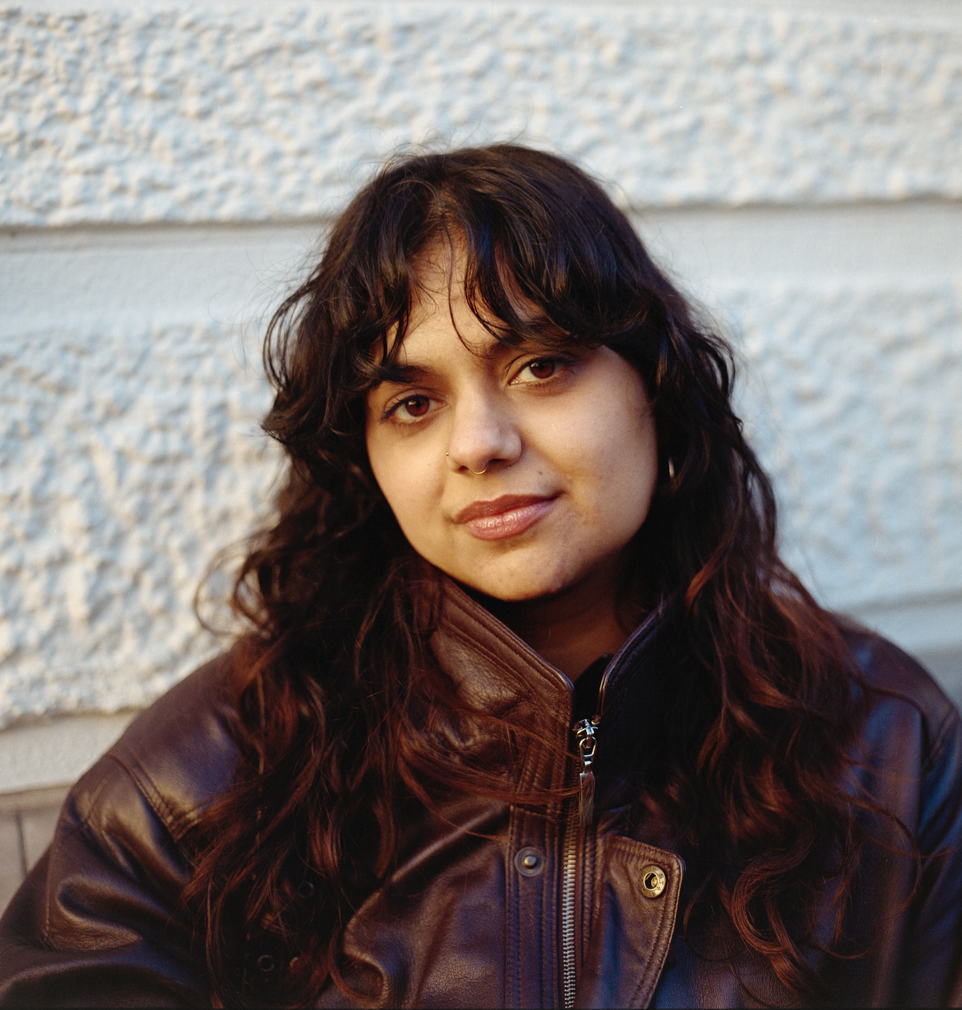 Close-up portrait of a woman with dark wavy hair, wearing a brown leather jacket, standing against a textured white wall.
