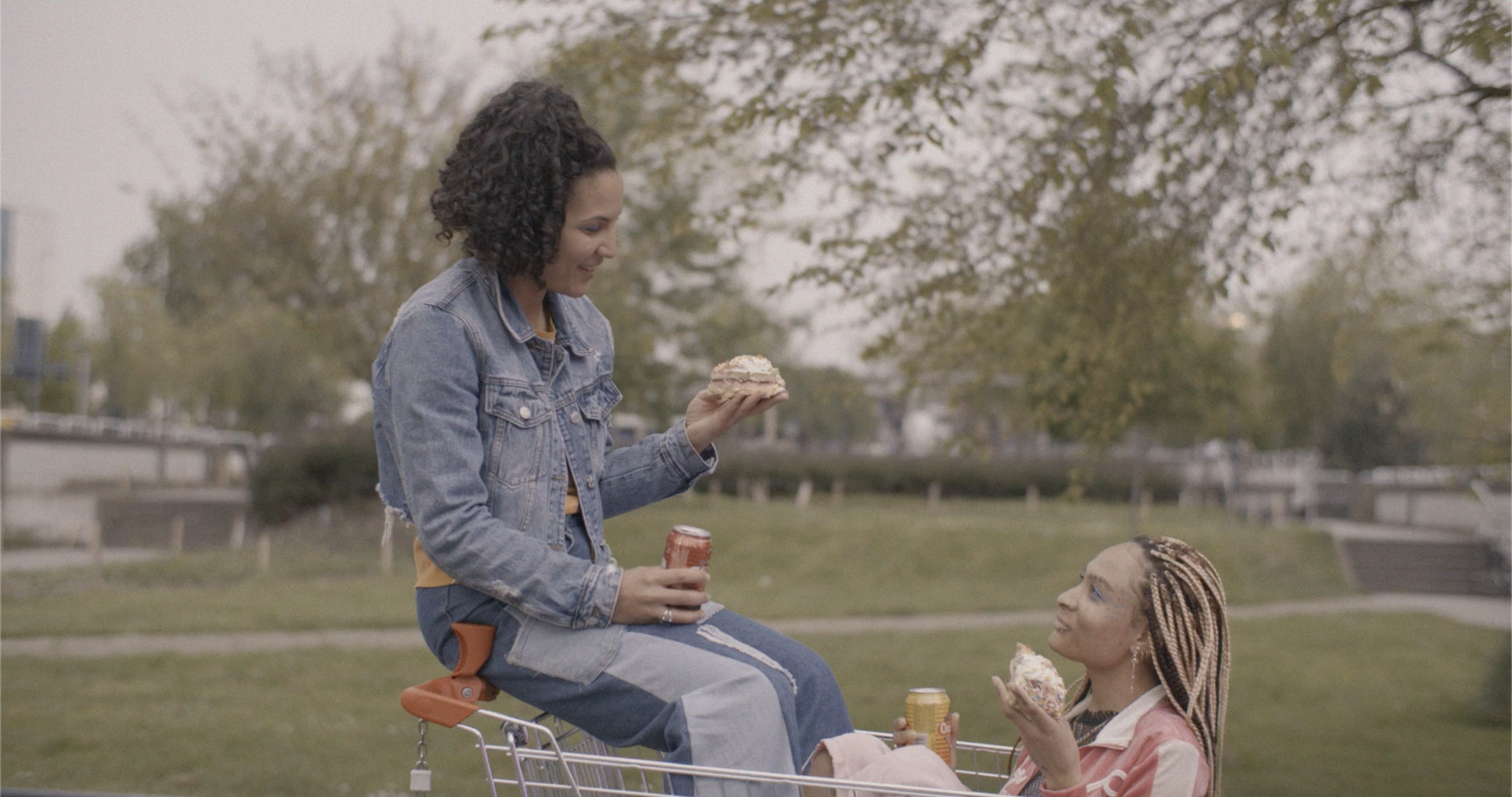 A woman with curly hair and a denim jacket sitting on a shopping cart, holding a soda can and a cupcake, and a girl with braided hair and face paint sitting inside the cart, holding a cupcake and a drink, outdoors in a park with trees in the backgrou
