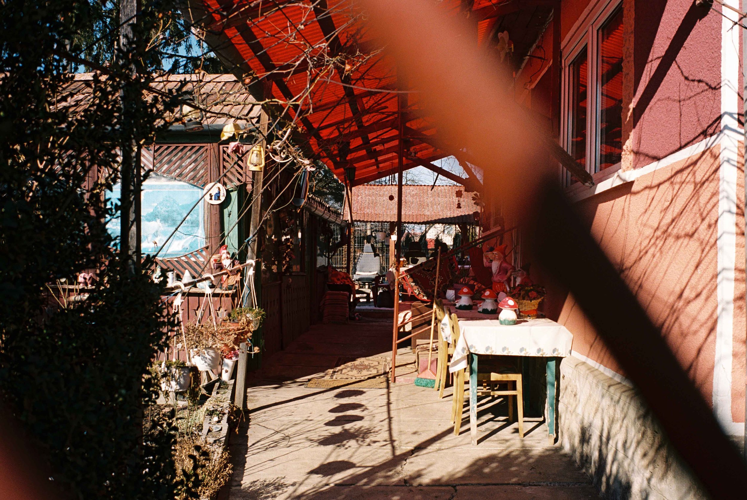 A cozy outdoor patio decorated with string lights, potted plants, and small garden gnomes, with a table covered by a tablecloth and surrounded by chairs, set against the backdrop of a reddish house with a screened porch and windows.