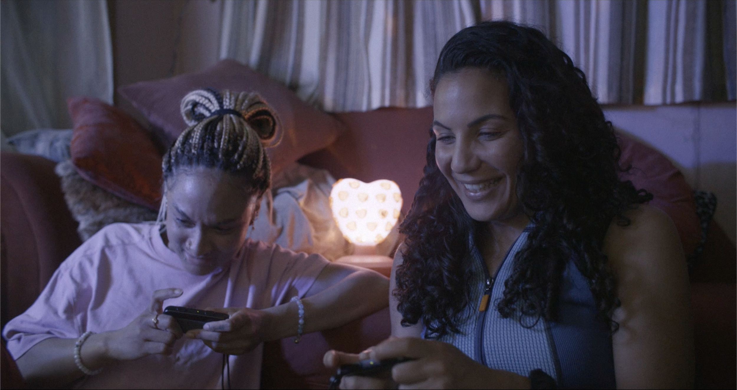 A woman with long curly hair smiling and a young girl with braided hair looking at a mobile phone in a dimly lit room with a glowing heart-shaped lamp on the bedside table.