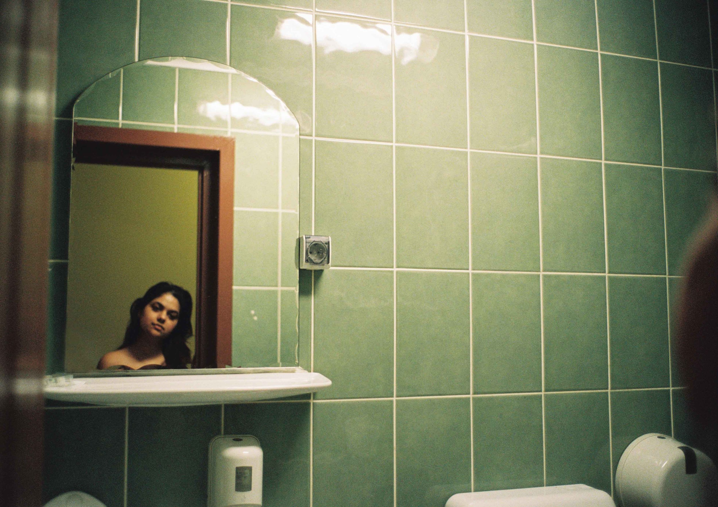 A woman with dark hair looking into a mirror in a bathroom with green tiled walls. The mirror reflects her face and part of the bathroom, including a soap dispenser and a toilet. Self portret