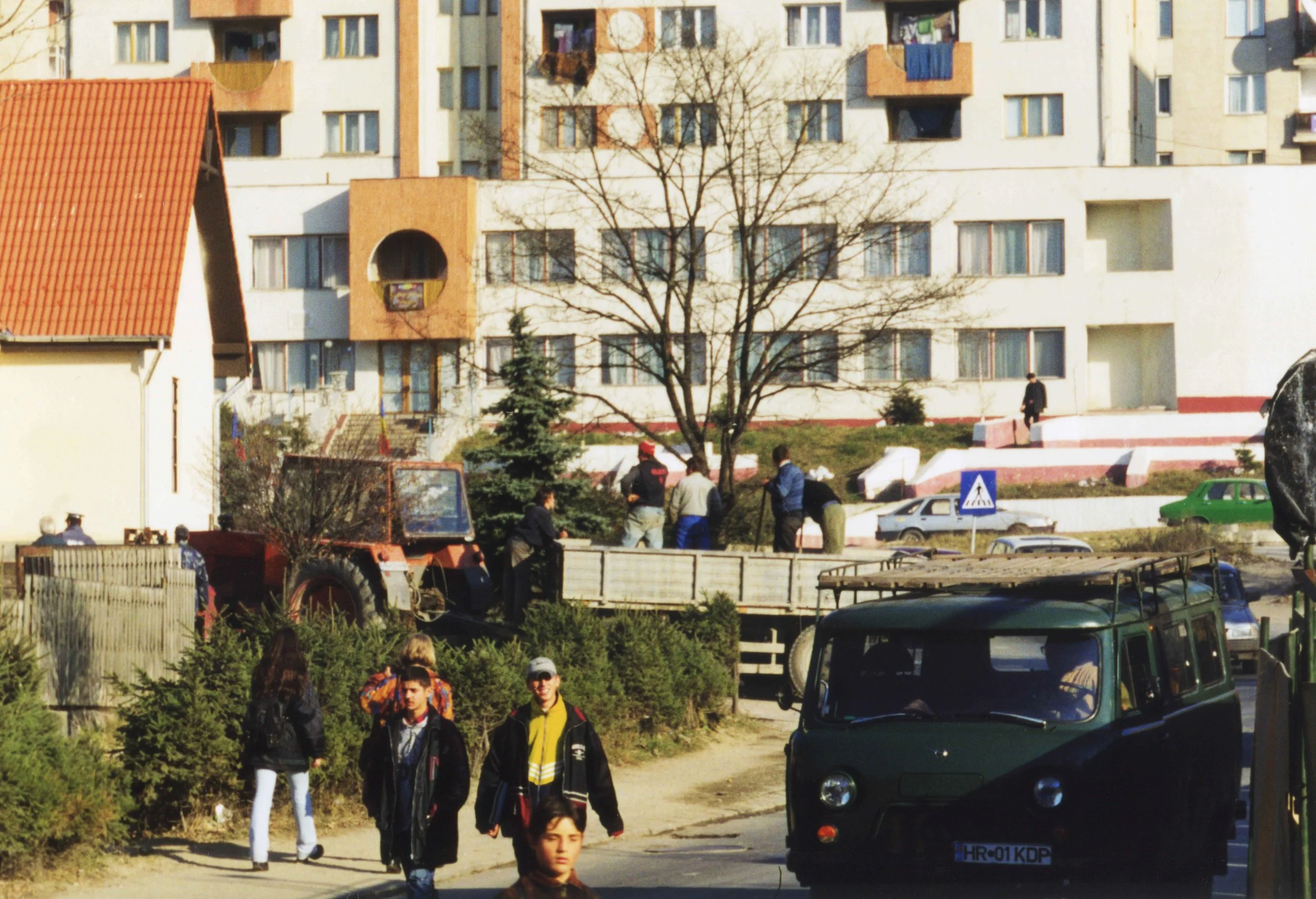 A residential urban scene with people walking, parked cars, and a large cream building with multiple balconies in the background. The image has a tree in the center and some small shrubs in the foreground. Romanian archive