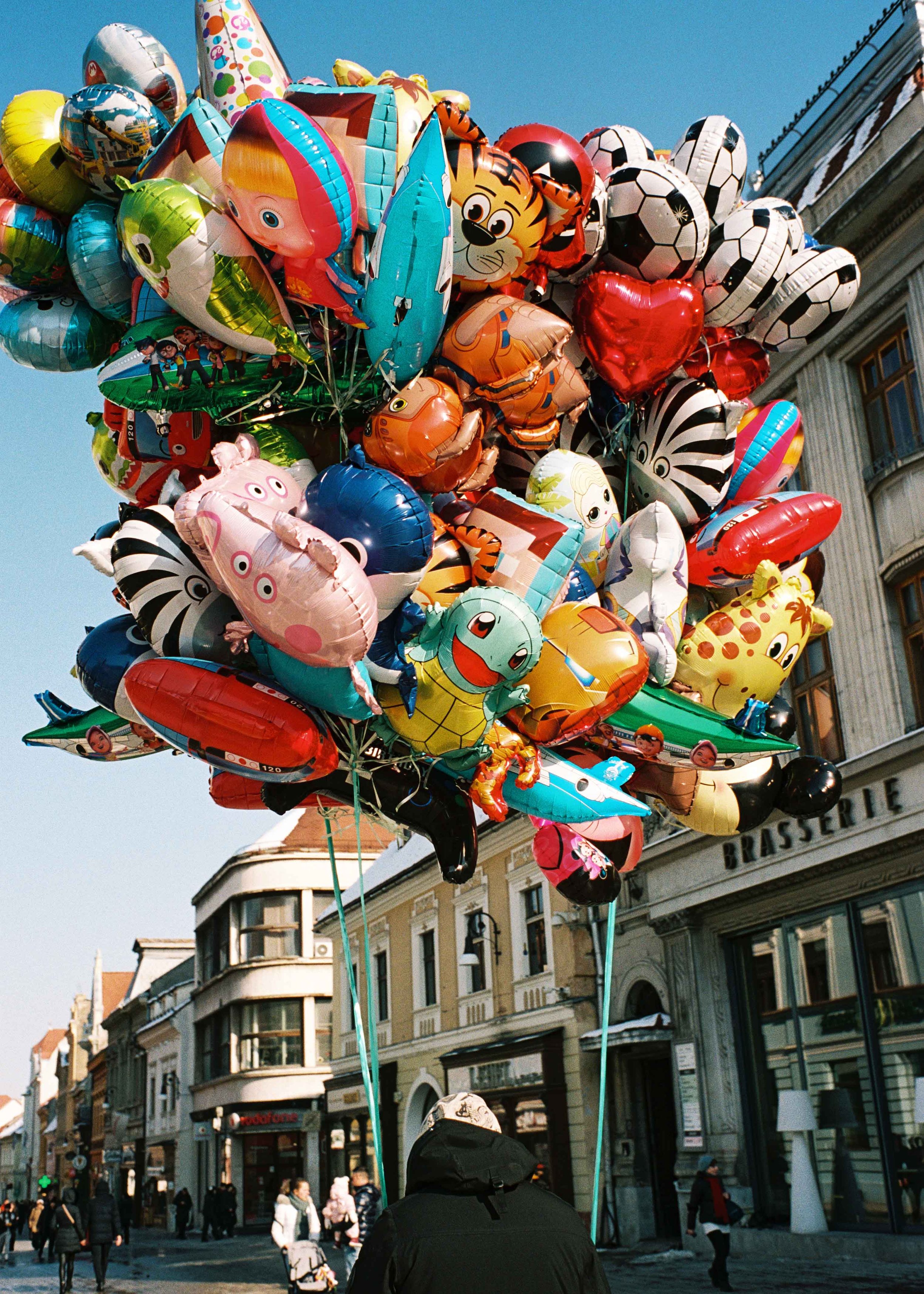 A person with a black jacket and a hat holding a bundle of colorful balloons shaped like animals, cartoon characters, and objects, in a busy city street with historic buildings and pedestrians.