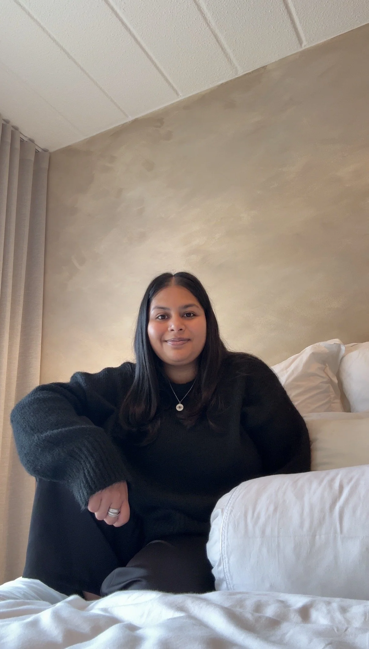 Prishaniee with long dark hair sitting on a bed, smiling, with a beige wall and curtains in the background.