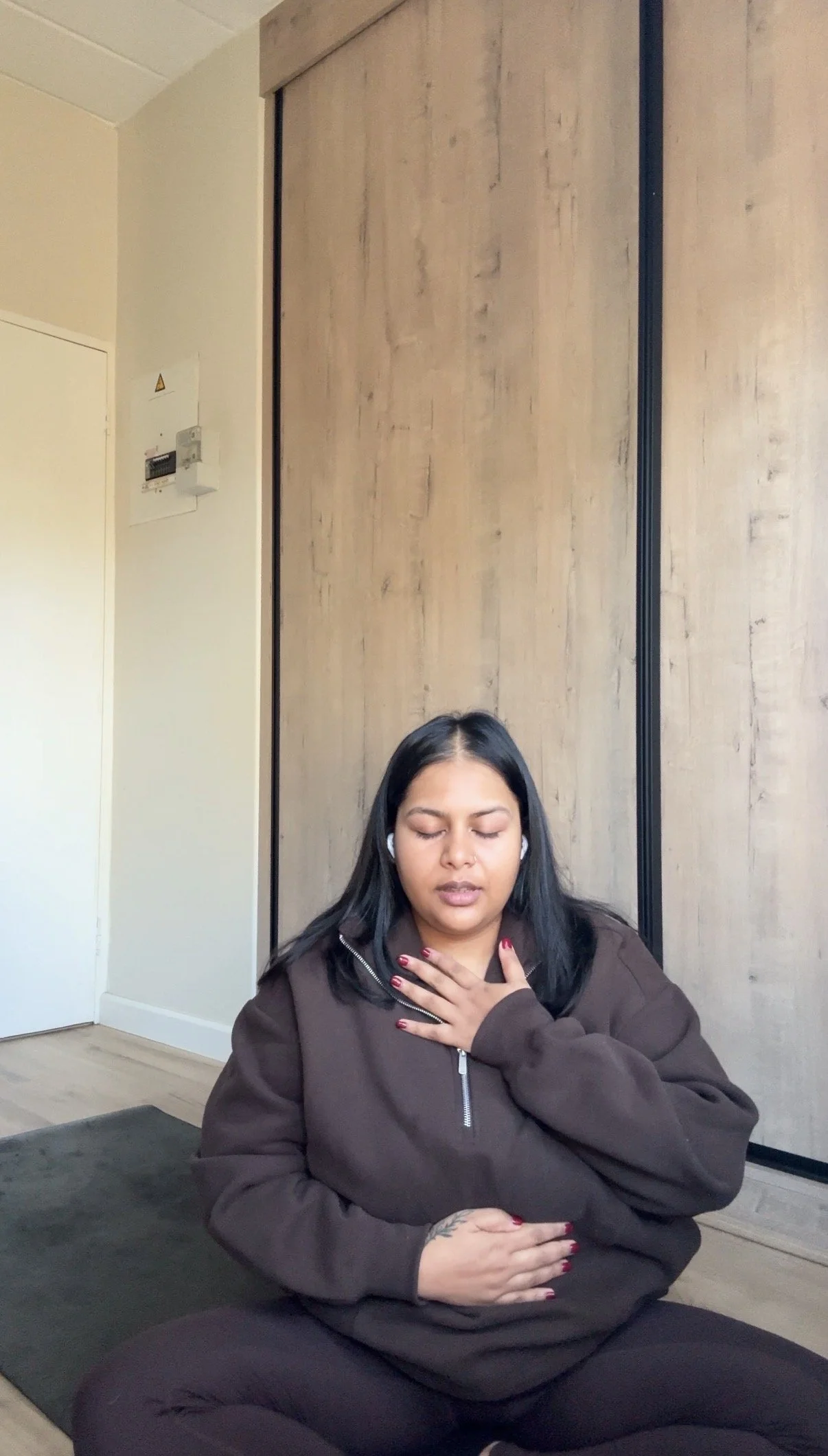 Prishaniee sitting cross-legged on a mat indoors with her eyes closed, one hand on her chest, and the other hand on her stomach, practicing meditation or mindfulness.