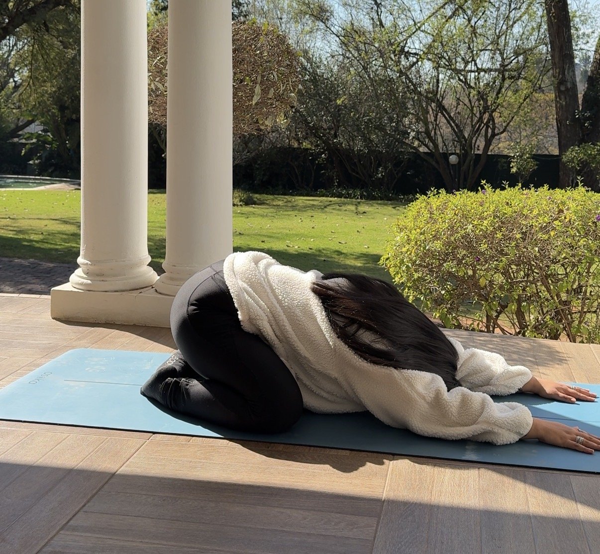 Prishaniee doing child's pose yoga stretch outside on a blue mat on a porch with white columns, green shrubs, and trees in the background.