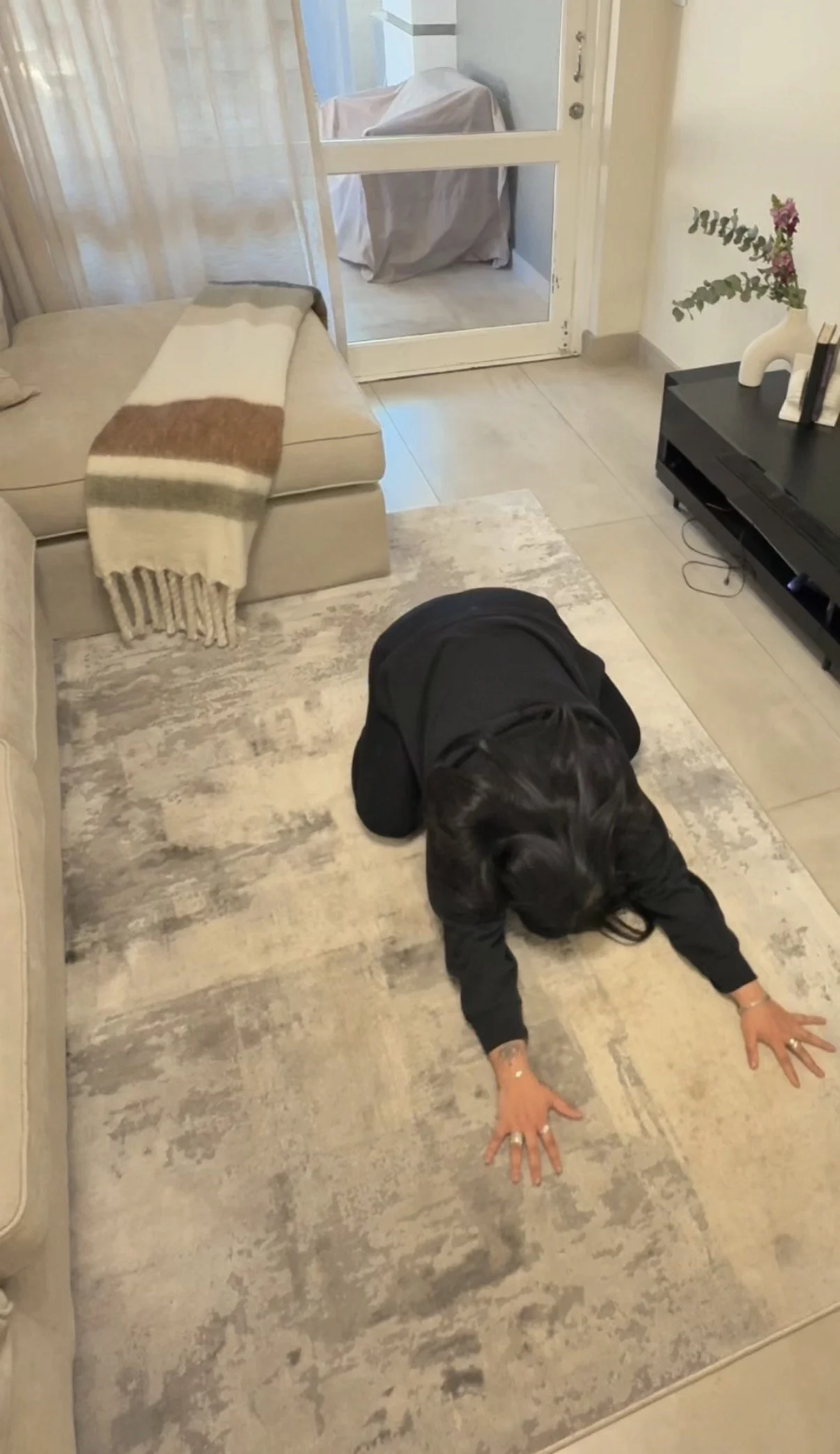 Prishaniee is on their hands and knees on a light-colored rug, in a room with a beige sofa, a throw blanket, a black TV stand with decor, and a glass door leading to a small enclosed porch or balcony.