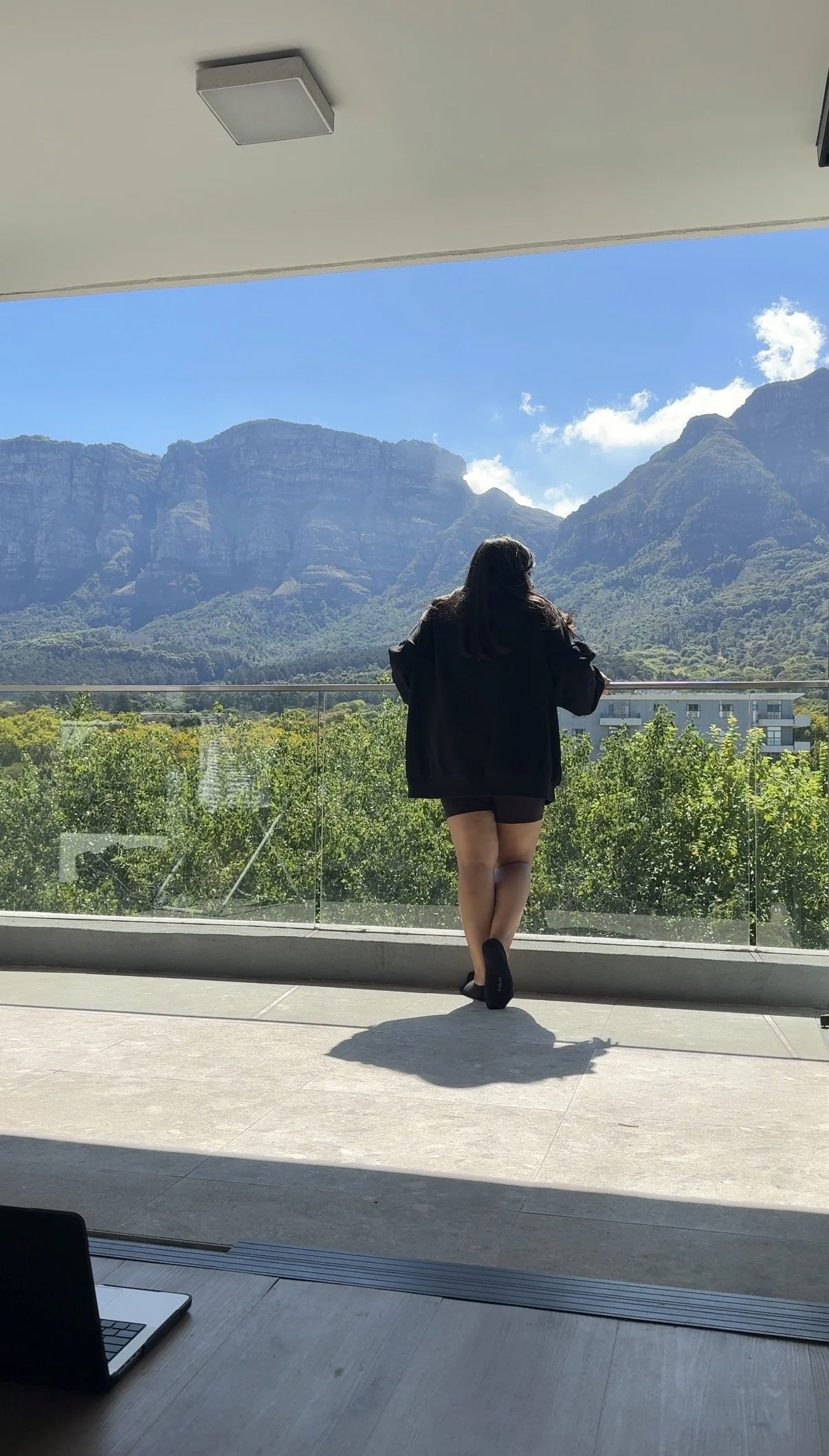 Prishaniee standing on a balcony looking at mountains with a blue sky, trees, and apartment buildings in the background.