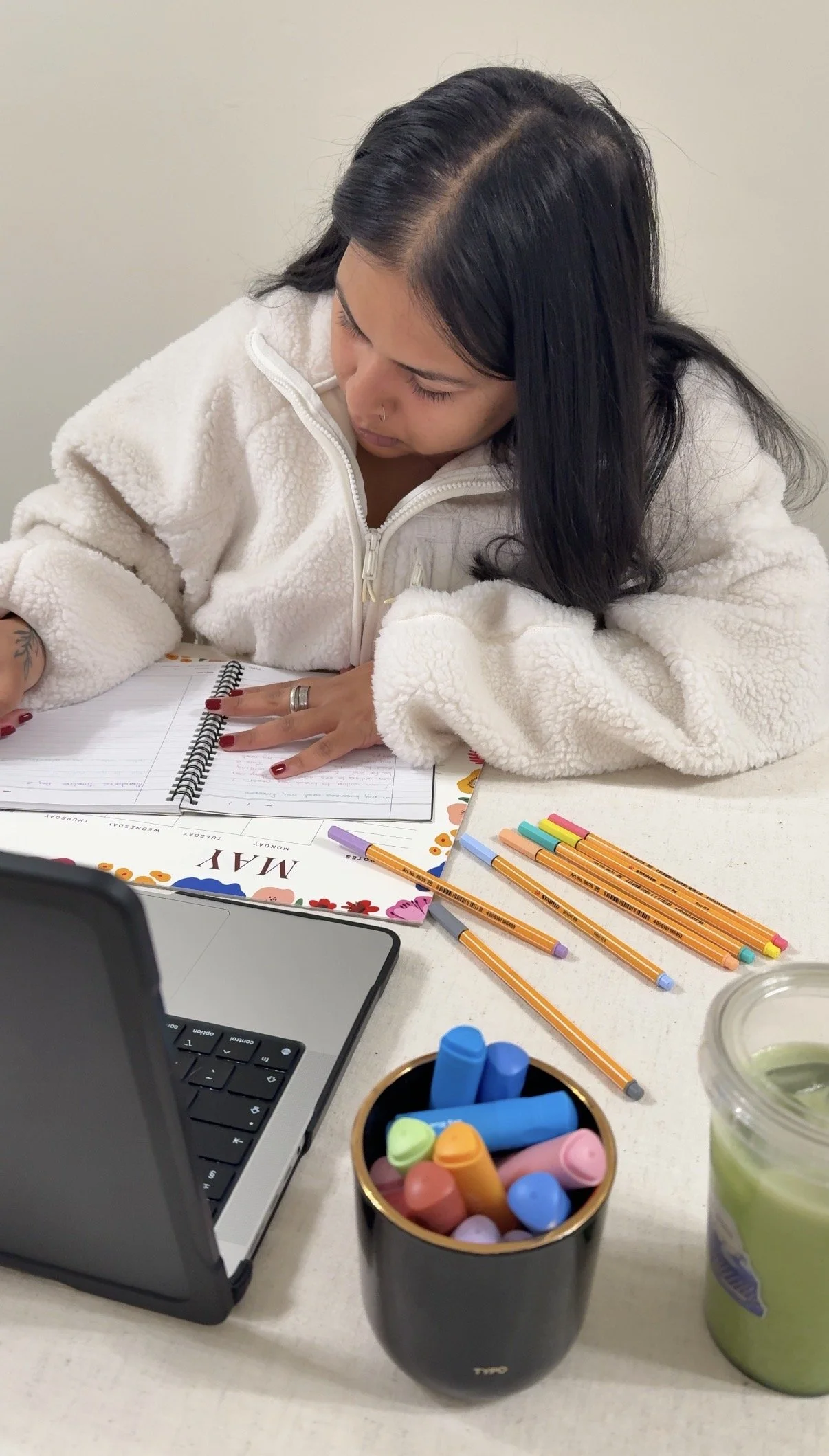 Prishaniee sitting at a table, writing in a planner with colorful highlighters and pens nearby, with a laptop on the table.