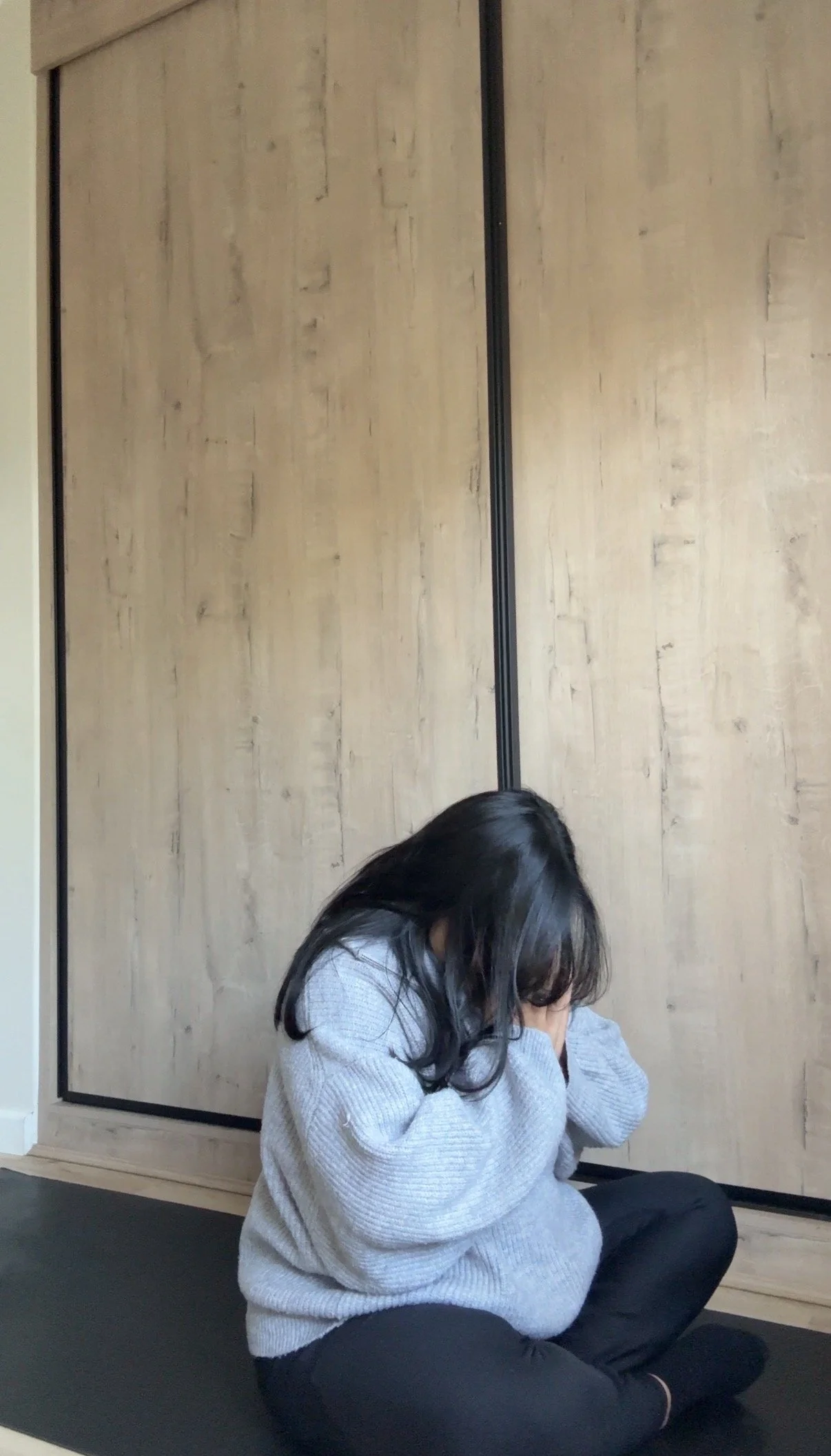 Prishaniee with long dark hair sitting cross-legged on a black yoga mat, wearing a gray sweater and black pants, with her hands covering her face, in front of a wooden wall.
