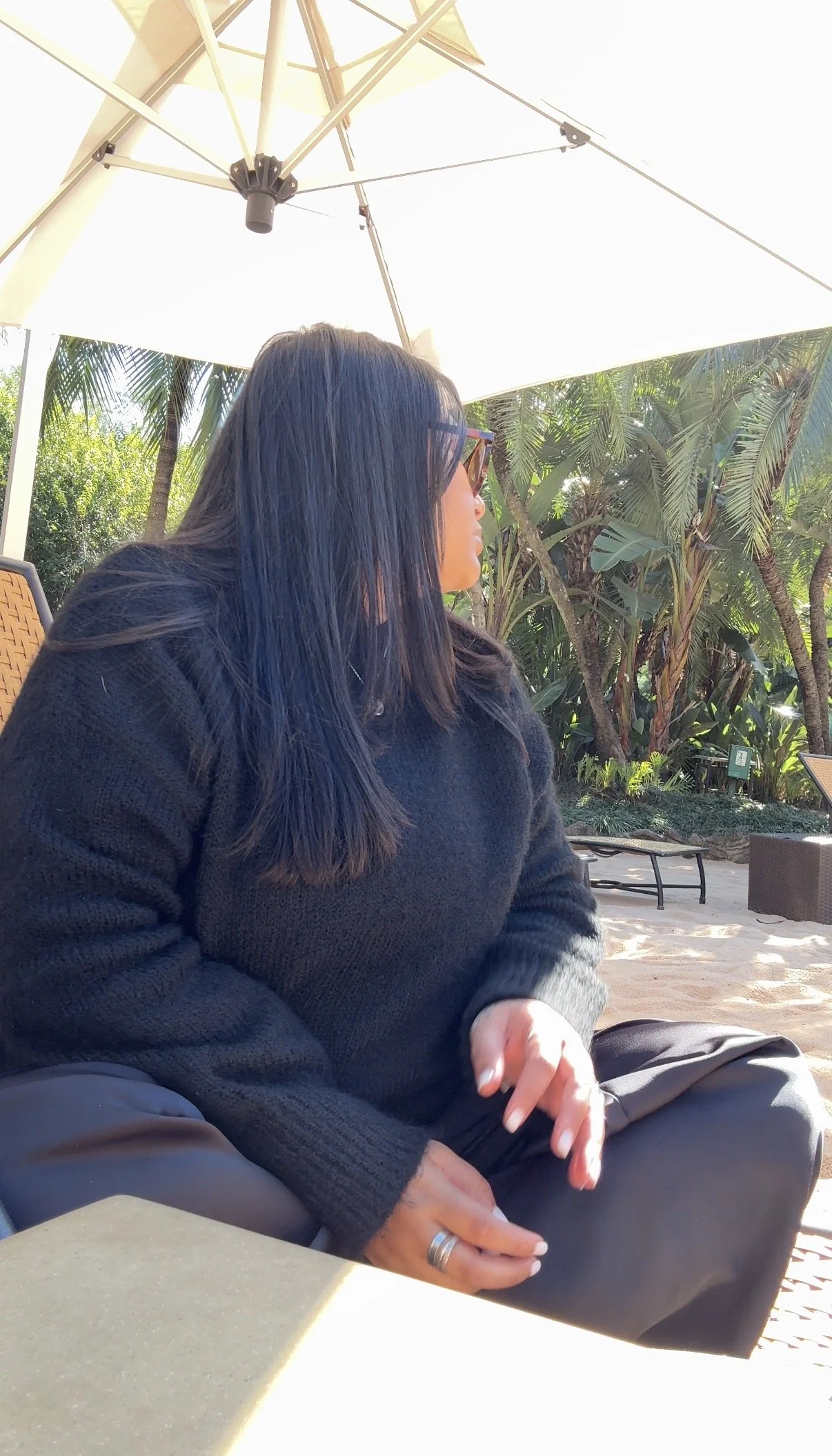 Prishaniee with long dark hair, wearing sunglasses and a black sweater, sitting outdoors under a large white patio umbrella surrounded by greenery and palm trees.