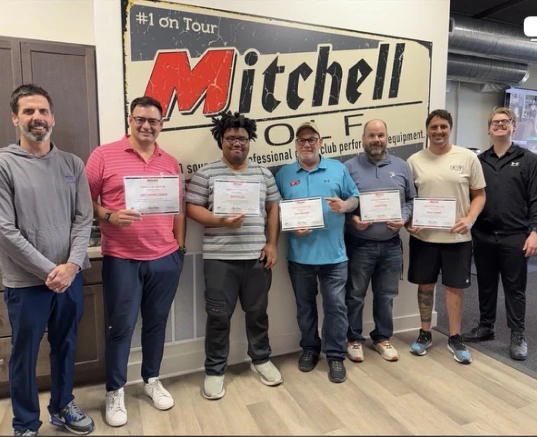 Group of seven men standing in front of a sign that reads "Mitchell Golf, #1 on Tour". Several men are holding certificates, and they are smiling. The setting appears to be a golf club or putting facility.