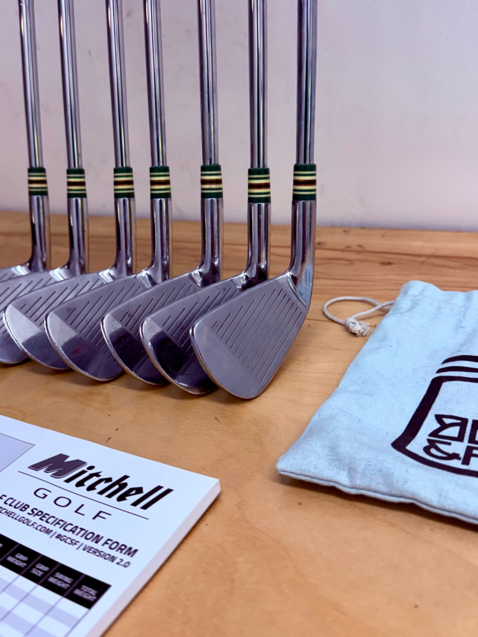 Six golf clubs lined up on a wooden table with a Mitchell Golf club specification sheet and a cloth bag nearby.