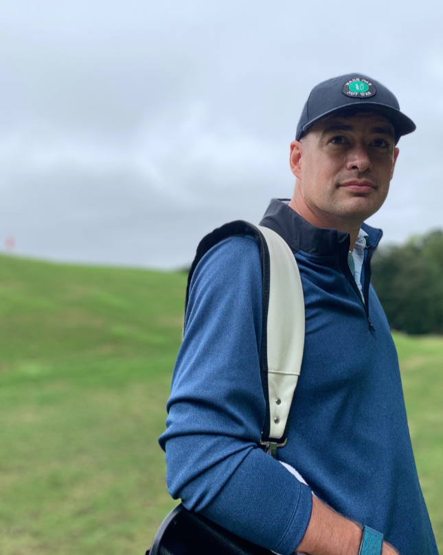 A man in a blue long-sleeve golf shirt and black cap with a green logo stands outdoors on a golf course with a grassy hill and cloudy sky in the background.