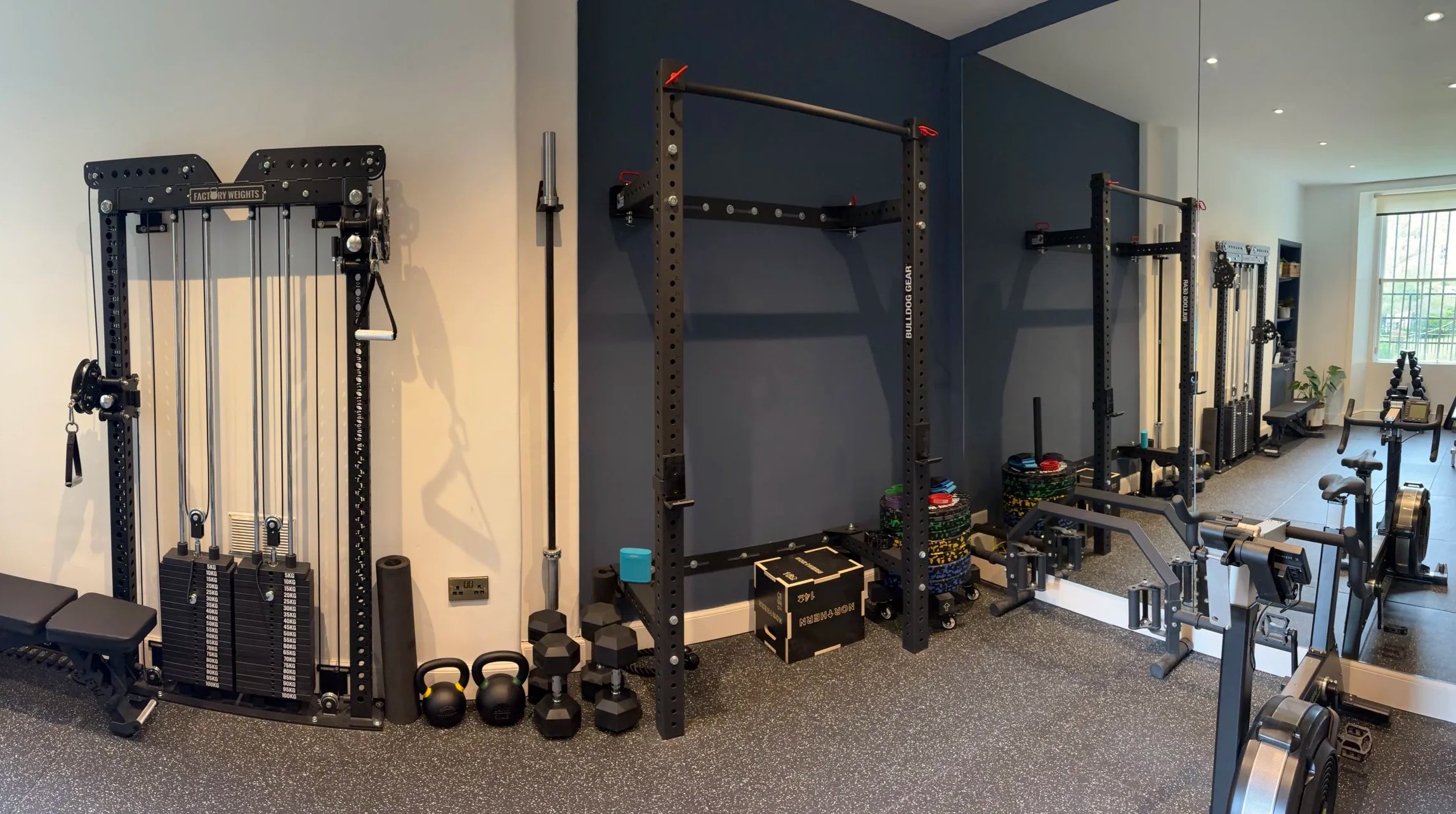 Home gym with black and gray workout equipment, including a weight machine with weights, kettlebells, dumbbells, resistance bands, a plyometric box, a rowing machine, and a mirror wall in a well-lit room with a window.