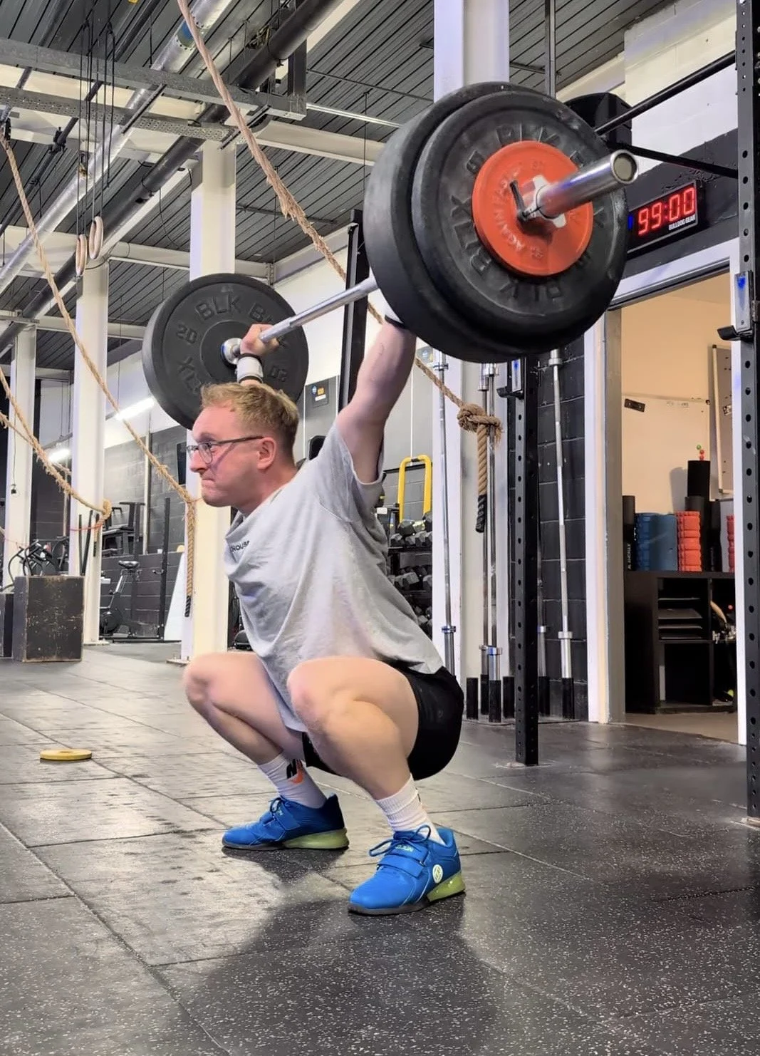 A man in a gym is performing a squat with a barbell loaded with weights, positioned overhead. He is wearing a gray t-shirt, black shorts, white socks, and blue sneakers. The gym has a scoreboard displaying 95:00, and various workout equipment are visible in the background.