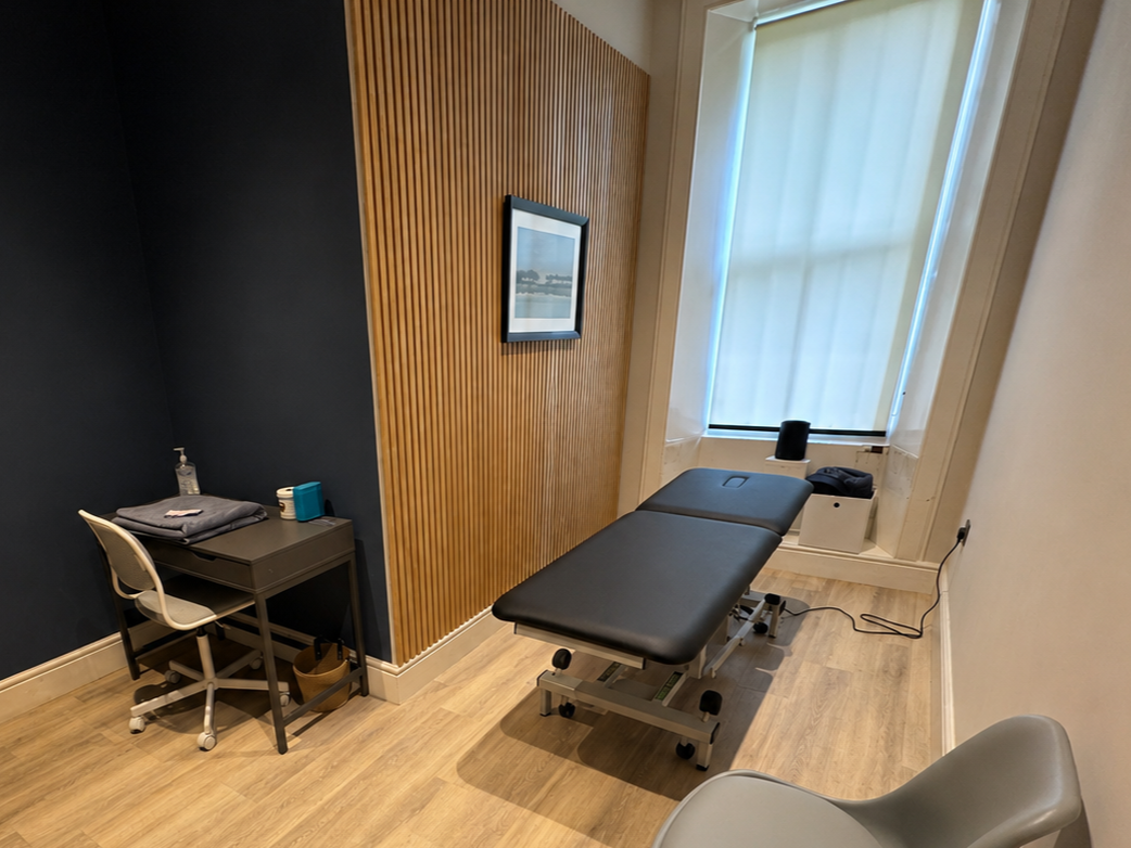 A medical examination room with a black treatment table by a large window with blinds, a small desk with medical supplies, and a chair.