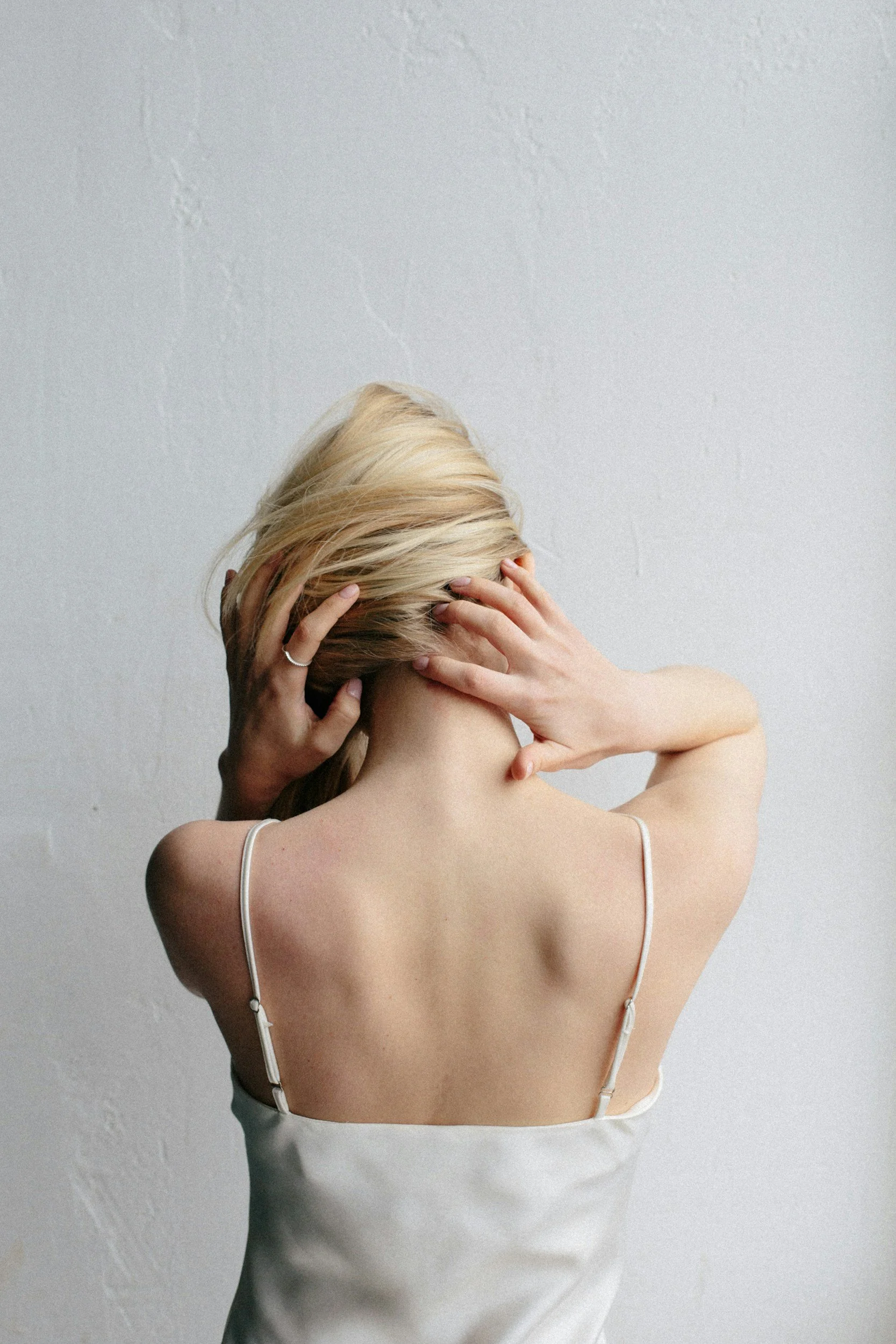 A woman with blonde hair wearing a white spaghetti strap dress, holding her head with both hands against a plain textured wall.