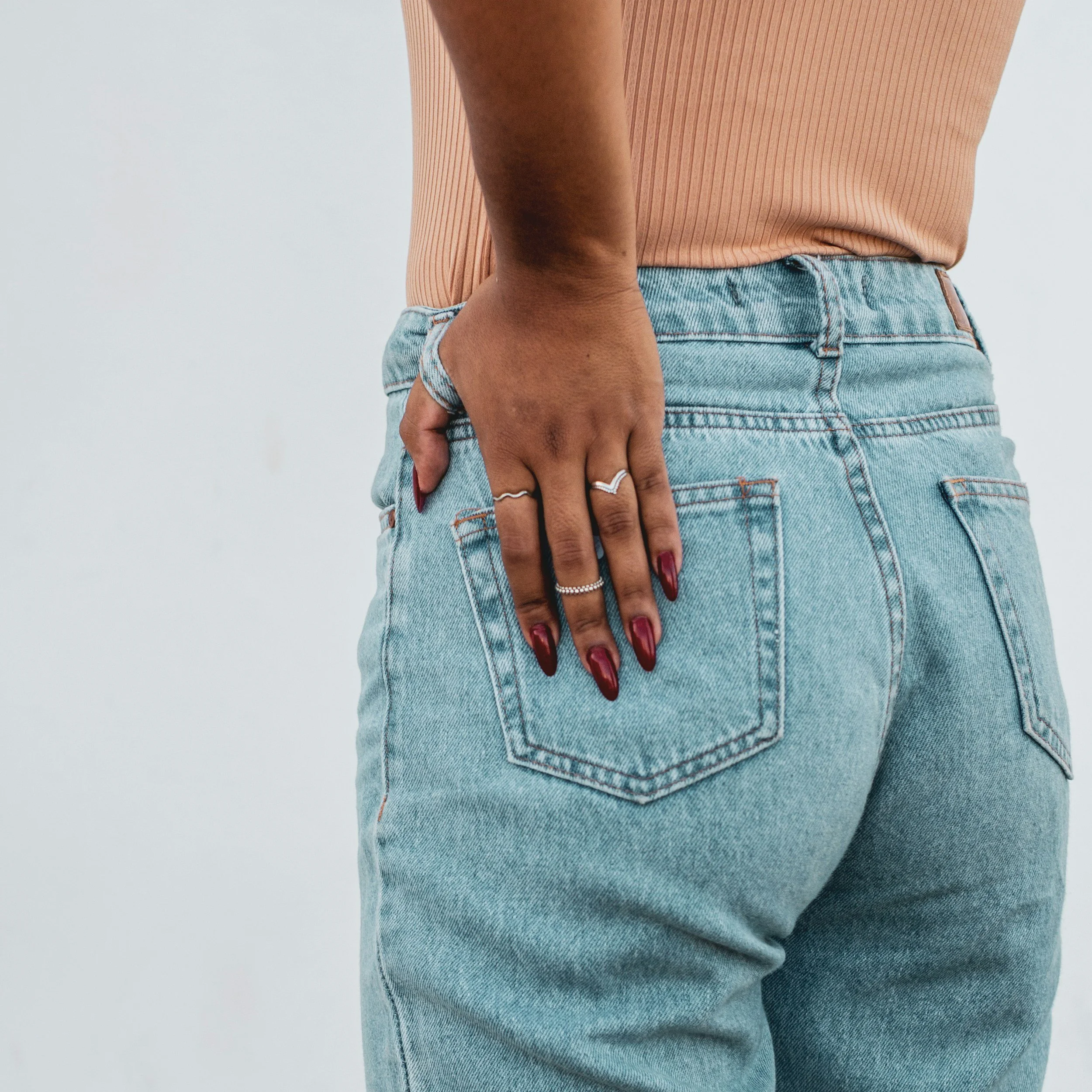 Close-up of a woman's hand resting on her hip, wearing a pink ribbed top and light-wash denim jeans against a plain background.