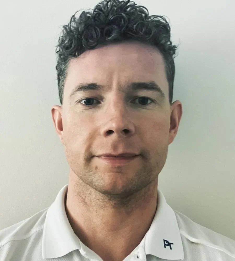 Headshot of a man with curly dark hair, wearing a white collared shirt with a small logo, standing against a plain light-colored wall.