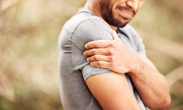 A man in a gray shirt is holding his left shoulder with his right hand, outdoors with a blurred natural background.