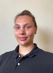 A young woman with brown hair tied back, wearing a dark polo shirt, smiling softly against a plain light-colored background.