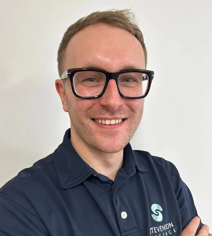 A smiling man with glasses and short light brown hair wearing a navy blue collared shirt with a logo on the chest, standing against a white background.