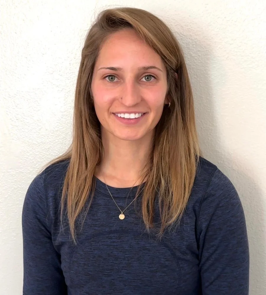 A young woman with long, light brown hair, blue eyes, and a nose piercing, smiling while standing against a plain white wall, wearing a dark blue top and a delicate necklace.