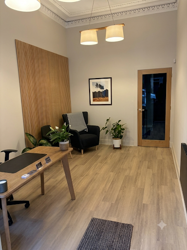 A modern waiting room with a black armchair, a small wooden side table, green plants, a framed picture on the wall, a wooden door with glass, and a wooden wall panel.