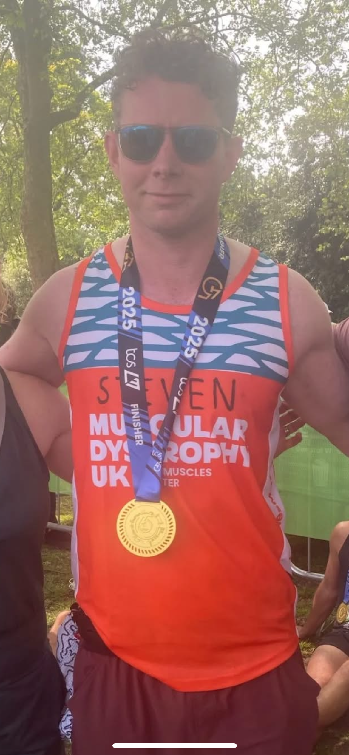 A man wearing sunglasses and a red and blue race shirt with a marathon or race medal around his neck, standing outdoors with trees in the background.