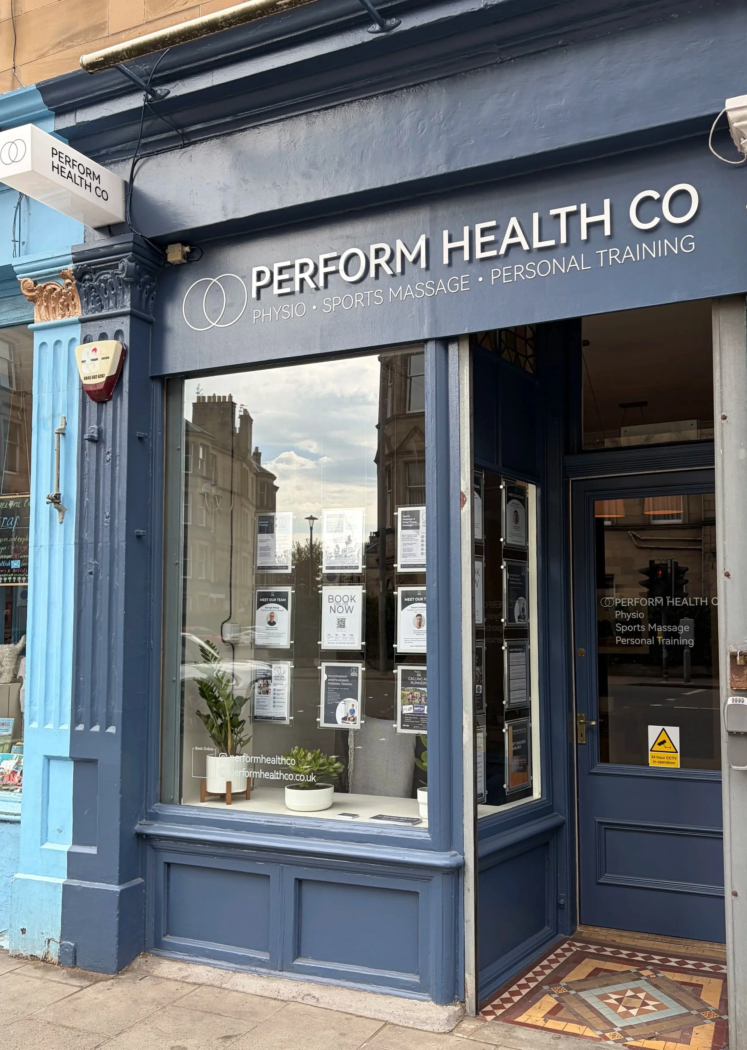 The storefront of Perform Health Co features a dark blue exterior with a large glass window displaying various posters and a small plant. The signage includes the company name and services such as physiotherapy, sports massage, and personal training. There are decorative architectural details around the window and door, with a colorful tiled floor at the entrance and a warning sign near the door.