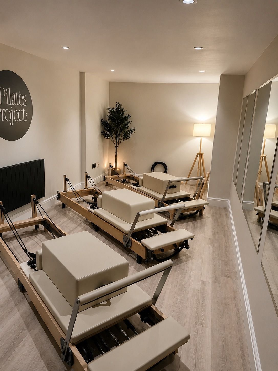 Pilates reformer machines in a fitness studio with mirrors on the wall, a floor lamp, a potted plant, and wall art that says "Pilates Project:".