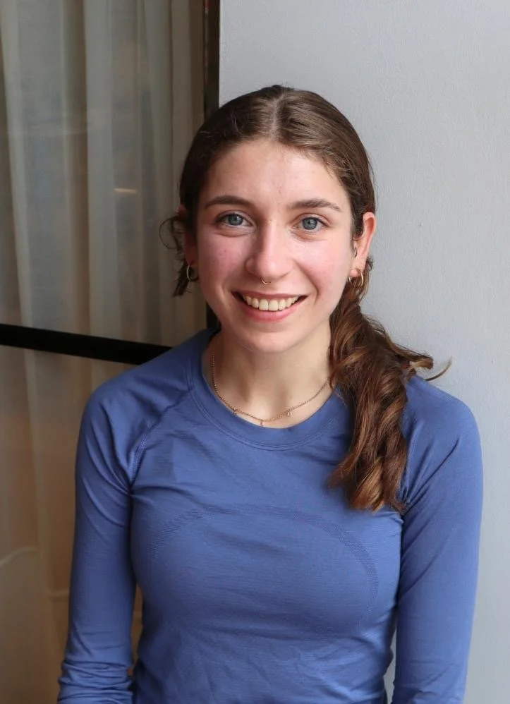 A young woman with brown hair tied to the side, wearing a blue long-sleeve shirt, smiling, standing indoors near a window and a white wall.