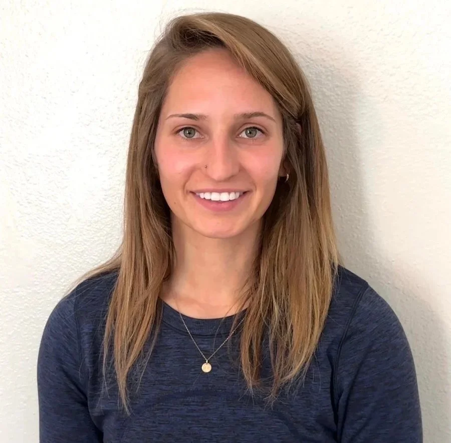 Portrait of a woman with light brown hair, smiling, wearing a navy blue shirt and a silver necklace, standing against a white wall.