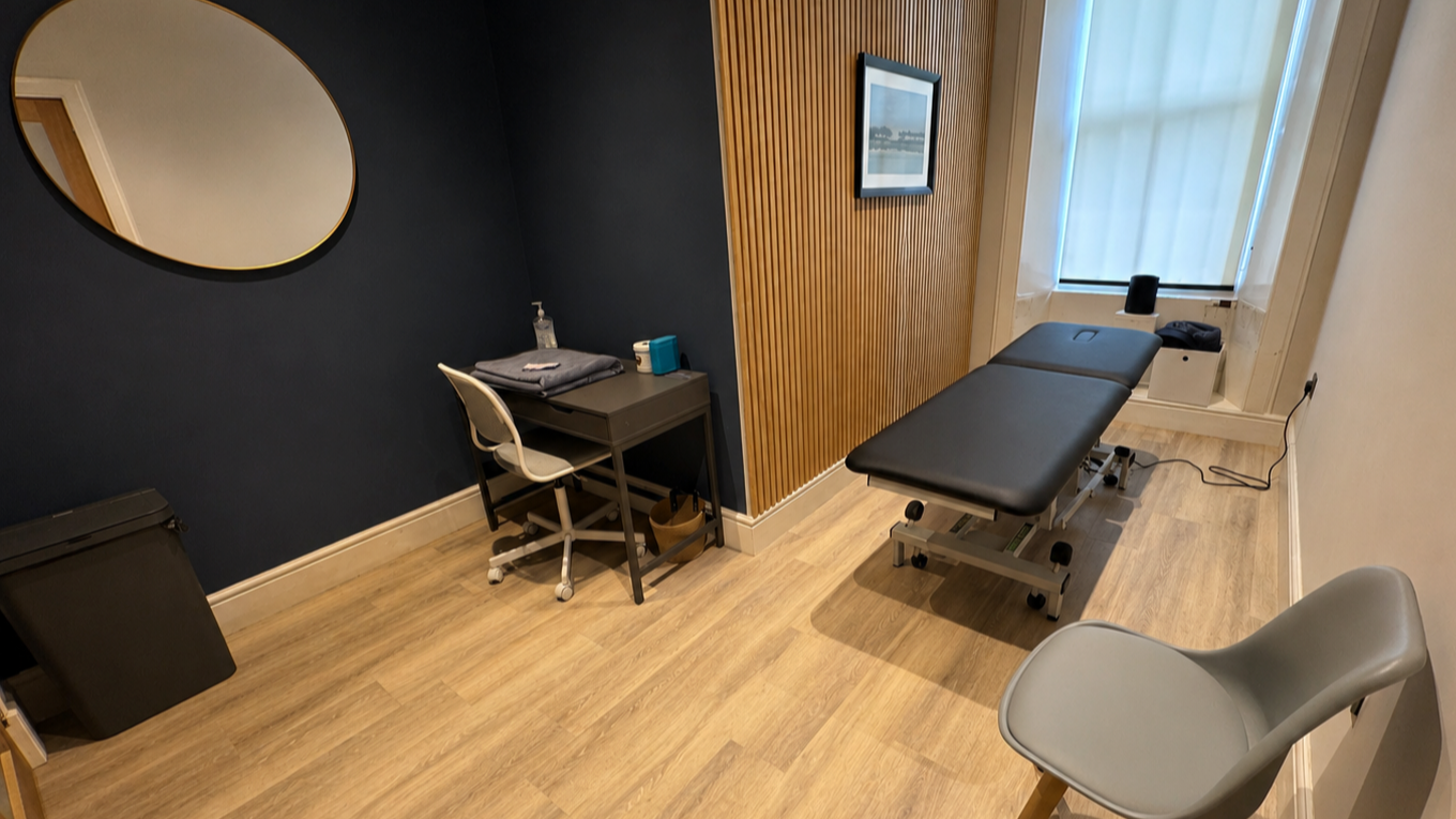 Empty medical or physiotherapy examination room with a treatment table, a small desk, a chair, and a wall-mounted mirror.