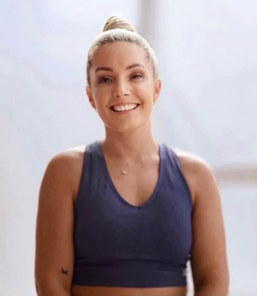 Smiling young woman with blonde hair in a bun, wearing a dark sports bra, in a bright indoor space.