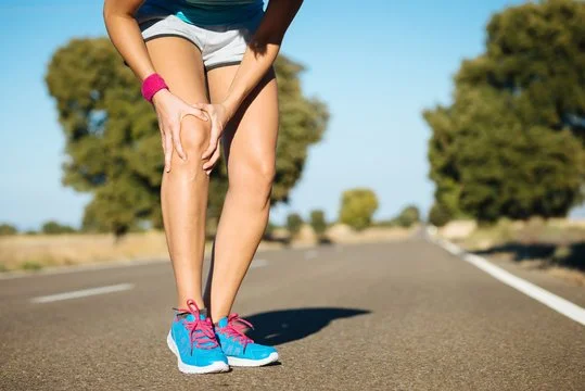 A woman is holding her knee on an empty road with trees in the background.