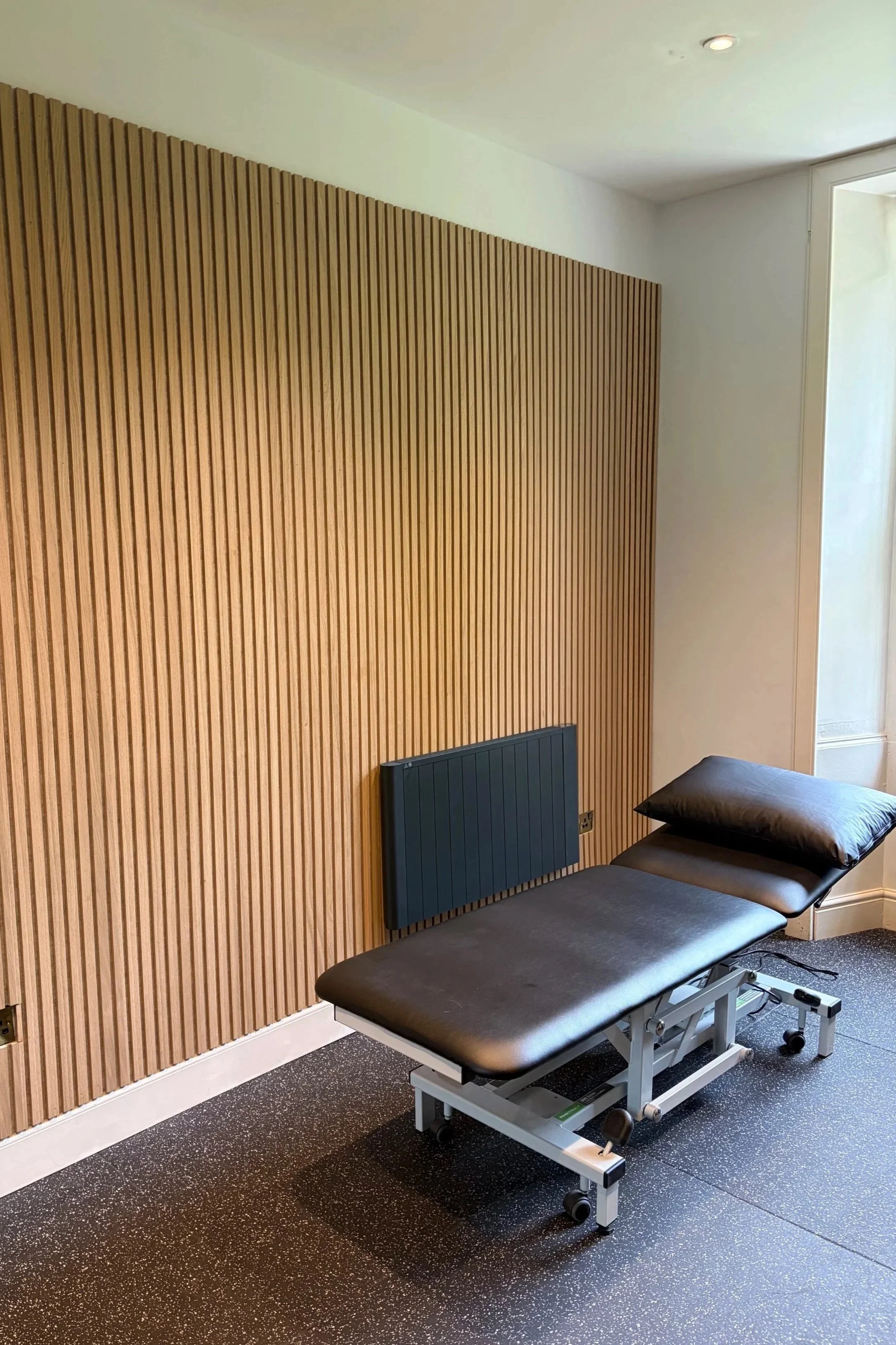 Medical examination room with a black examination bed with an adjustable headrest, pillows, wooden wall paneling, and a wall-mounted blue heater.