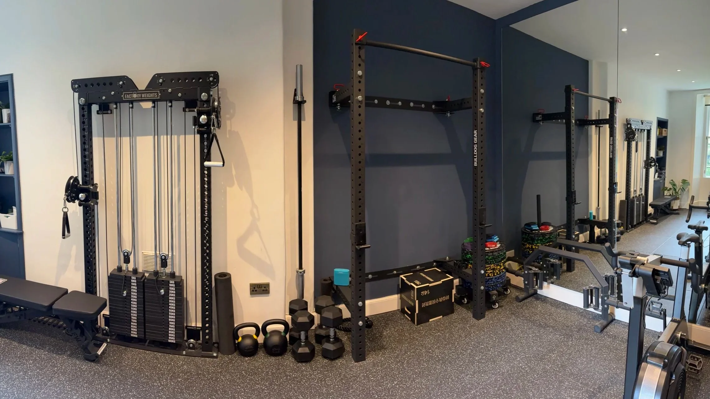 Home gym with various equipment including weight machine, dumbbells, kettlebells, plyometric boxes, and a squat rack, organized on a black rubber flooring with a mirror on the right wall.