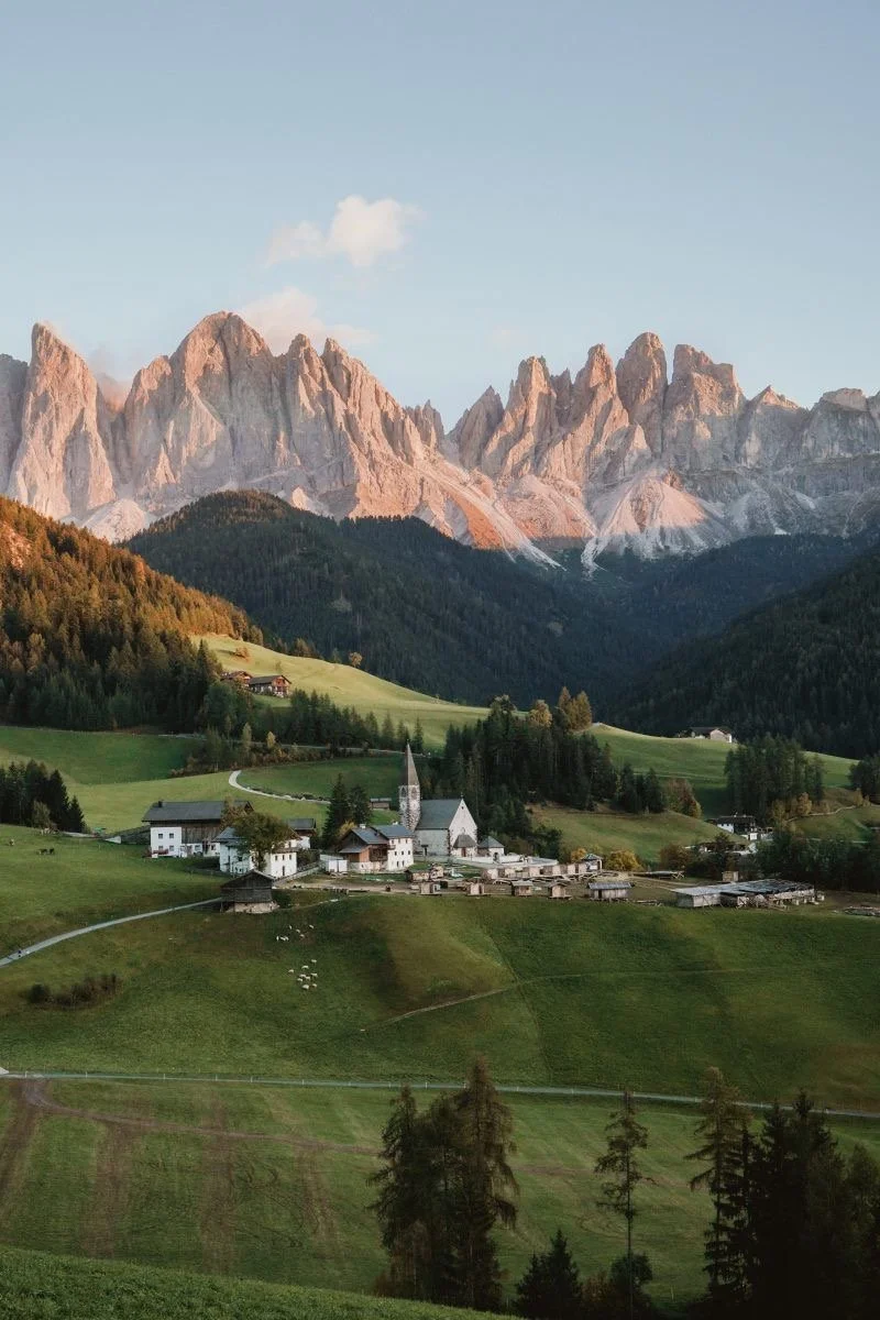 Scenic landscape of a small village with a church, green fields, and trees at the base of rugged, rocky mountains during sunset or sunrise.