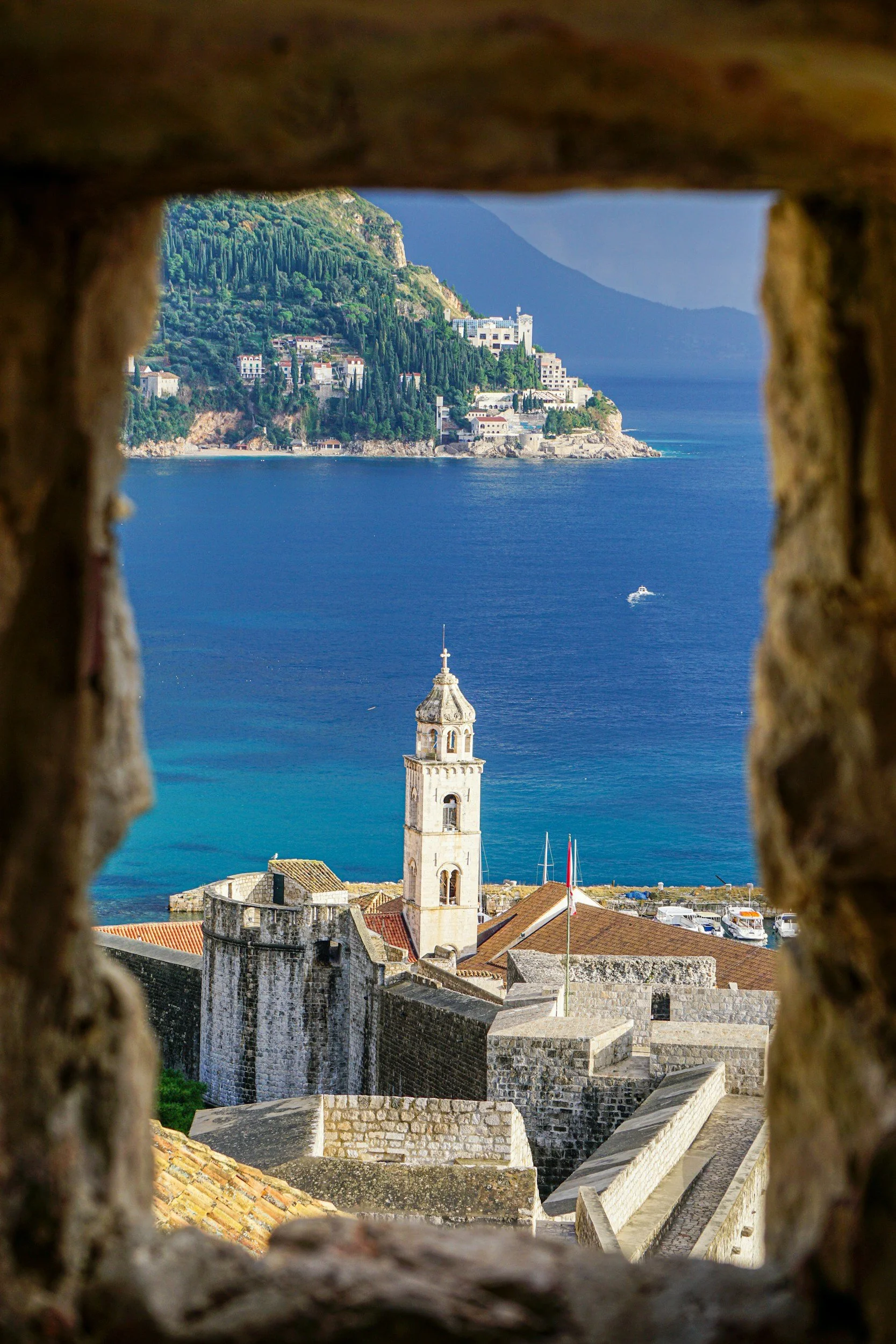 View of a coastal city with a stone tower and historic walls through the opening of a brick wall, overlooking the blue sea with a boat sailing nearby and a lush green hillside.
