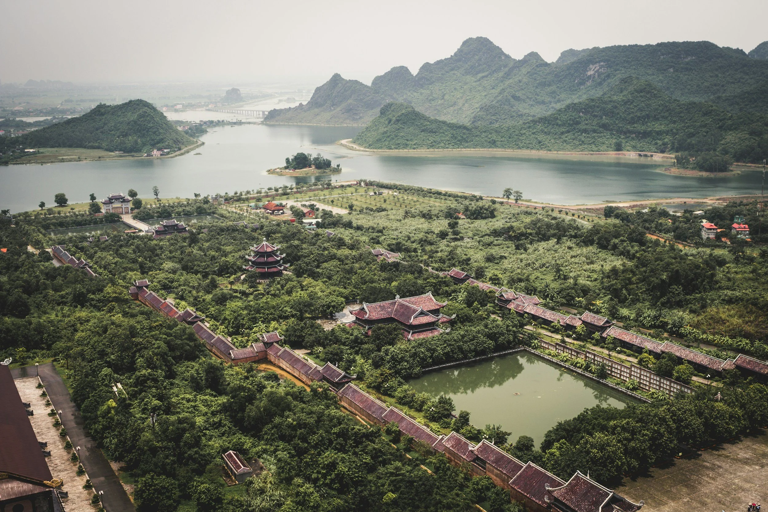 Scenic view of a lake surrounded by lush green hills and mountains, with traditional Asian-style buildings in the foreground and a bridge in the distance.