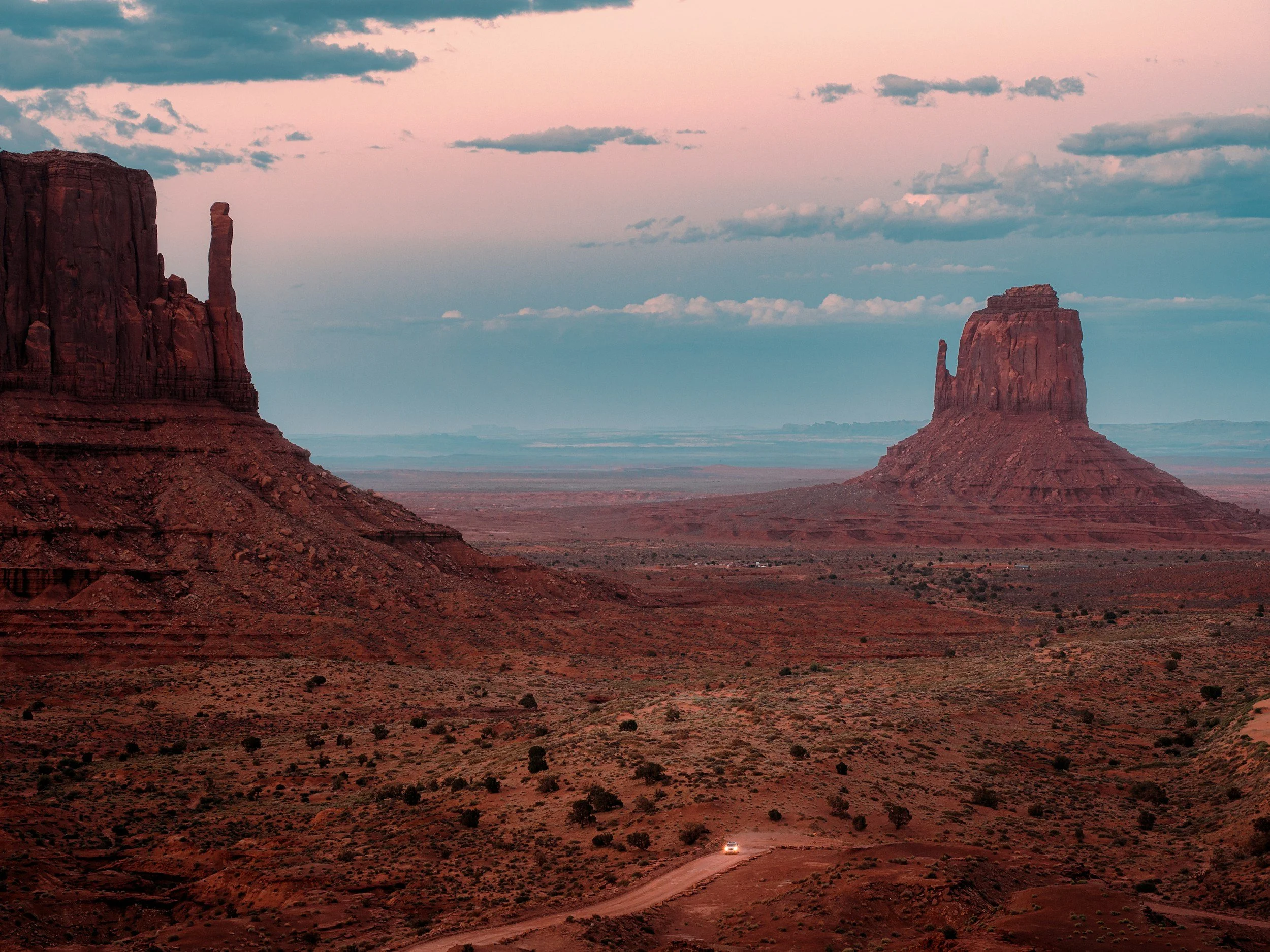 A vast desert landscape with large, red rock formations under a partly cloudy sky during sunset or sunrise. A small vehicle is visible on a dirt road at the bottom of the scene.