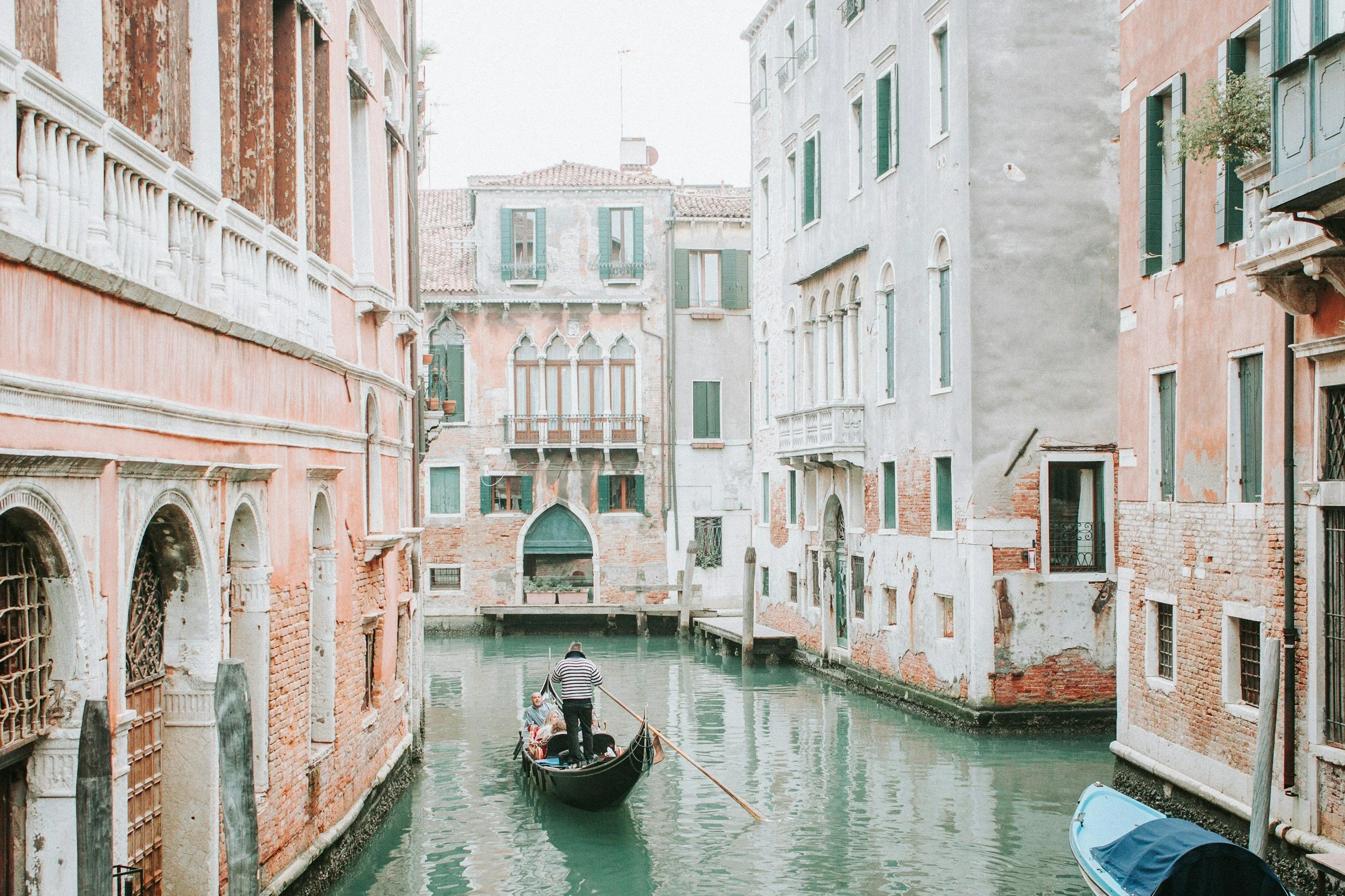 Gondola with passengers and gondolier navigating a canal in Venice, Italy, surrounded by historic buildings with colorful facades and green shutters.