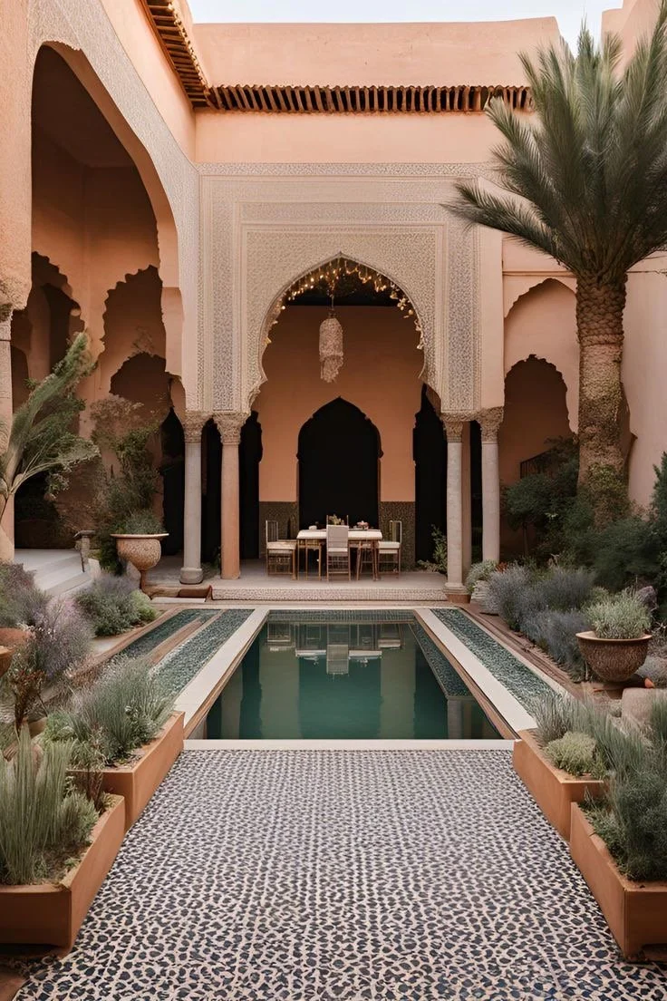 A courtyard with a swimming pool, Moroccan-style architecture, and lush desert plants, including palm trees. There is a table with chairs under a decorative archway, and the area is decorated with string lights and a chandelier.