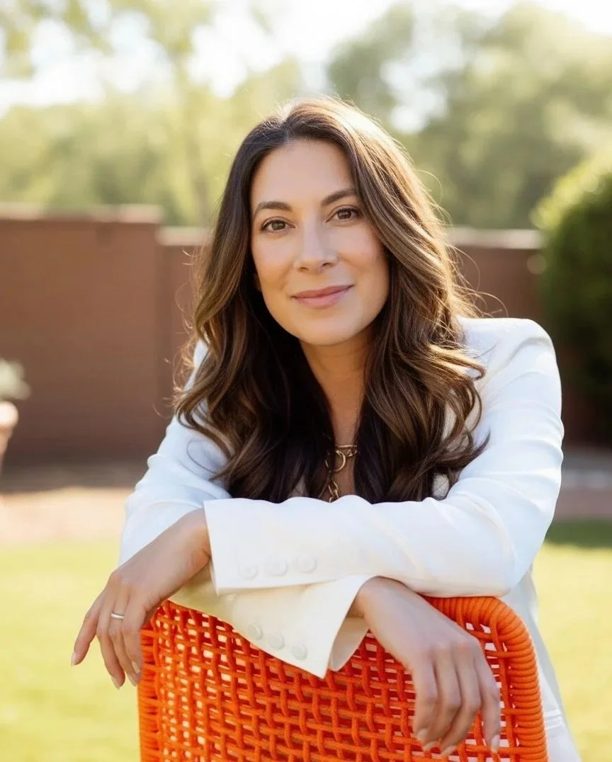 A woman sitting outdoors on an orange chair, smiling, with a blurred green background.