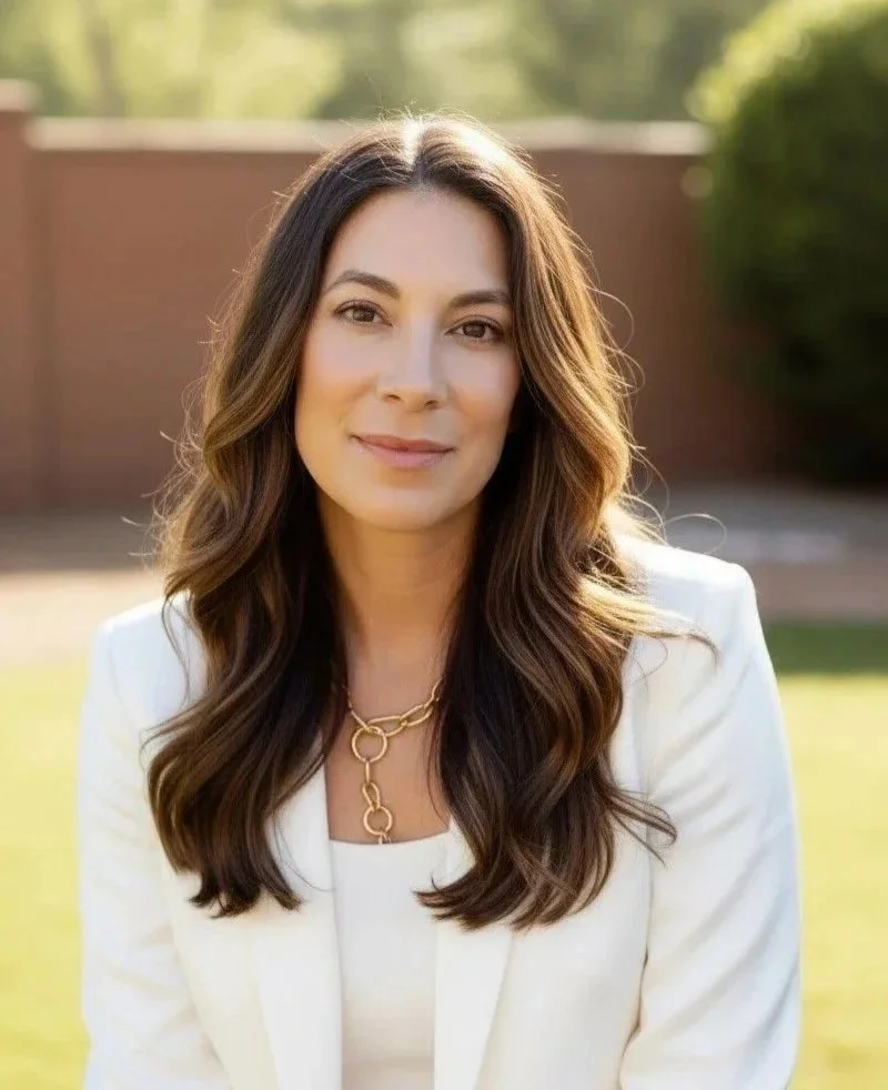 A woman with long wavy brown hair, wearing a white blazer and gold chain necklace, smiling outdoors with a blurred background of trees and a brick wall.