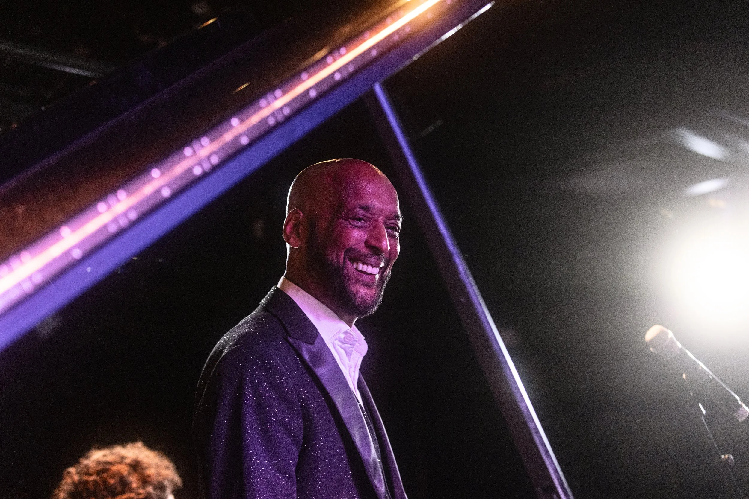 Tommy Blaize wearing a dark, shiny tuxedo, smiling at an event with dim lighting and colourful lights.