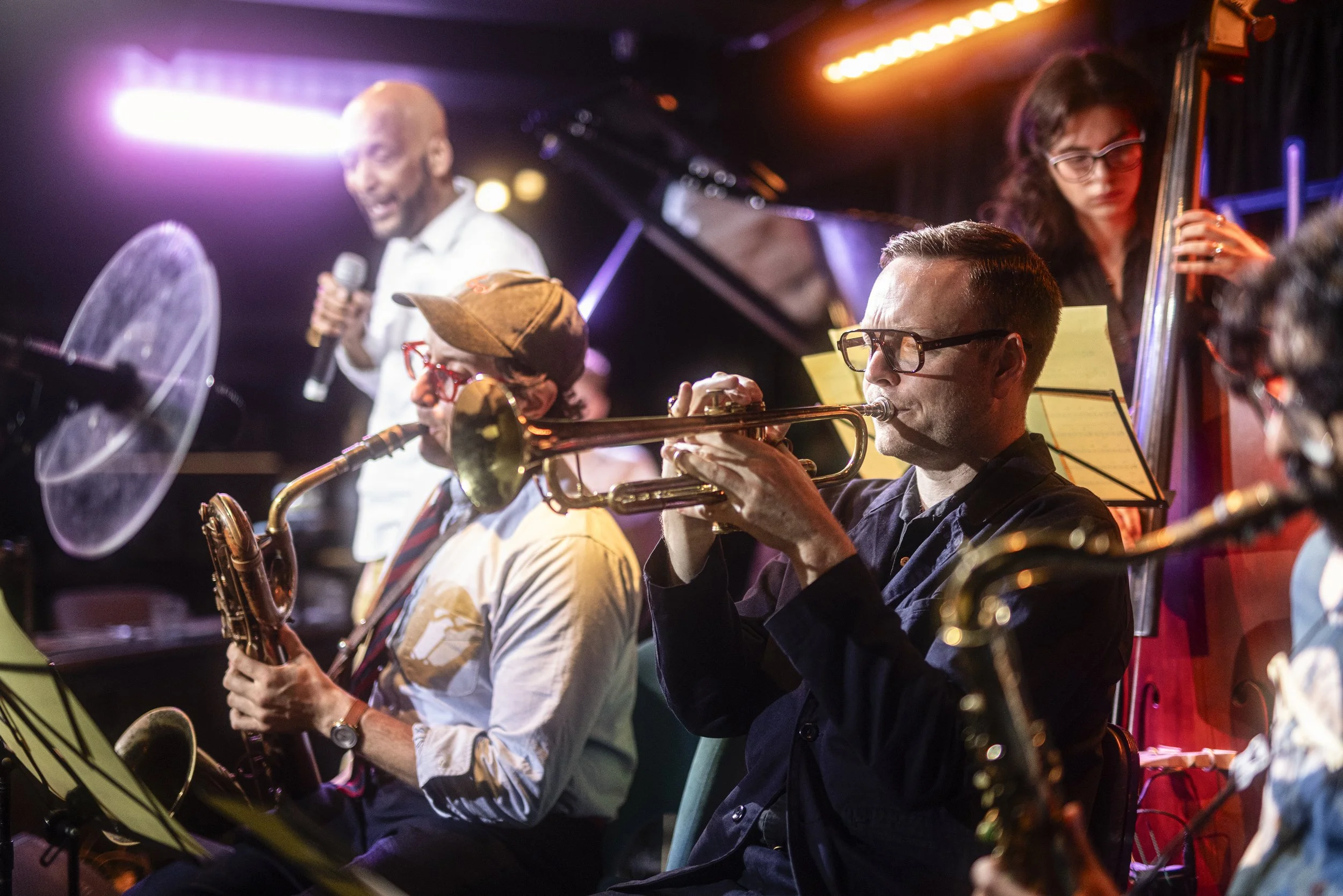 Jazz band performing on stage with Tommy Blaize holding a microphone, a saxophonist, and a double bass player, under colourful stage lighting.
